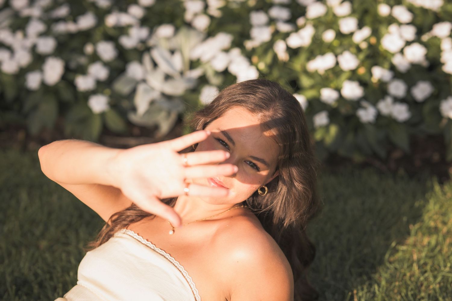 Young woman sitting on grass making peace sign, smiling in front of white blooming flowers in sunlight.