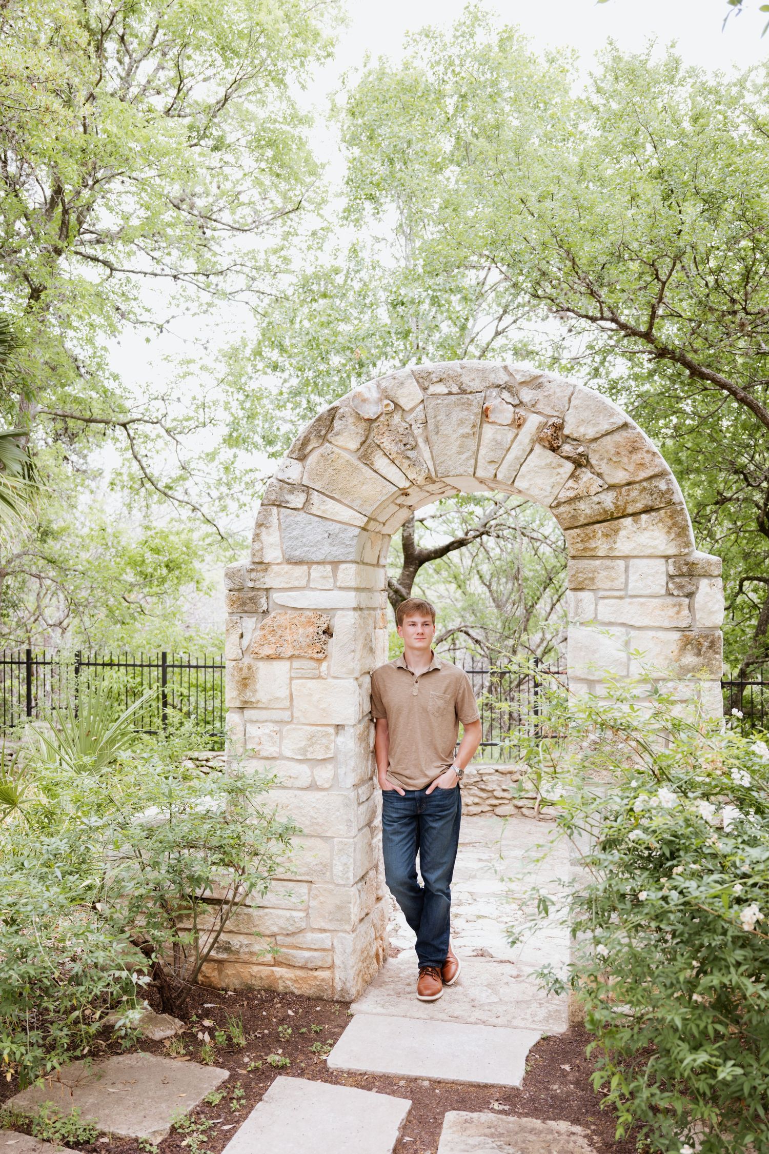 Young man posing under rustic stone garden arch surrounded by lush green trees in Austin, Texas.