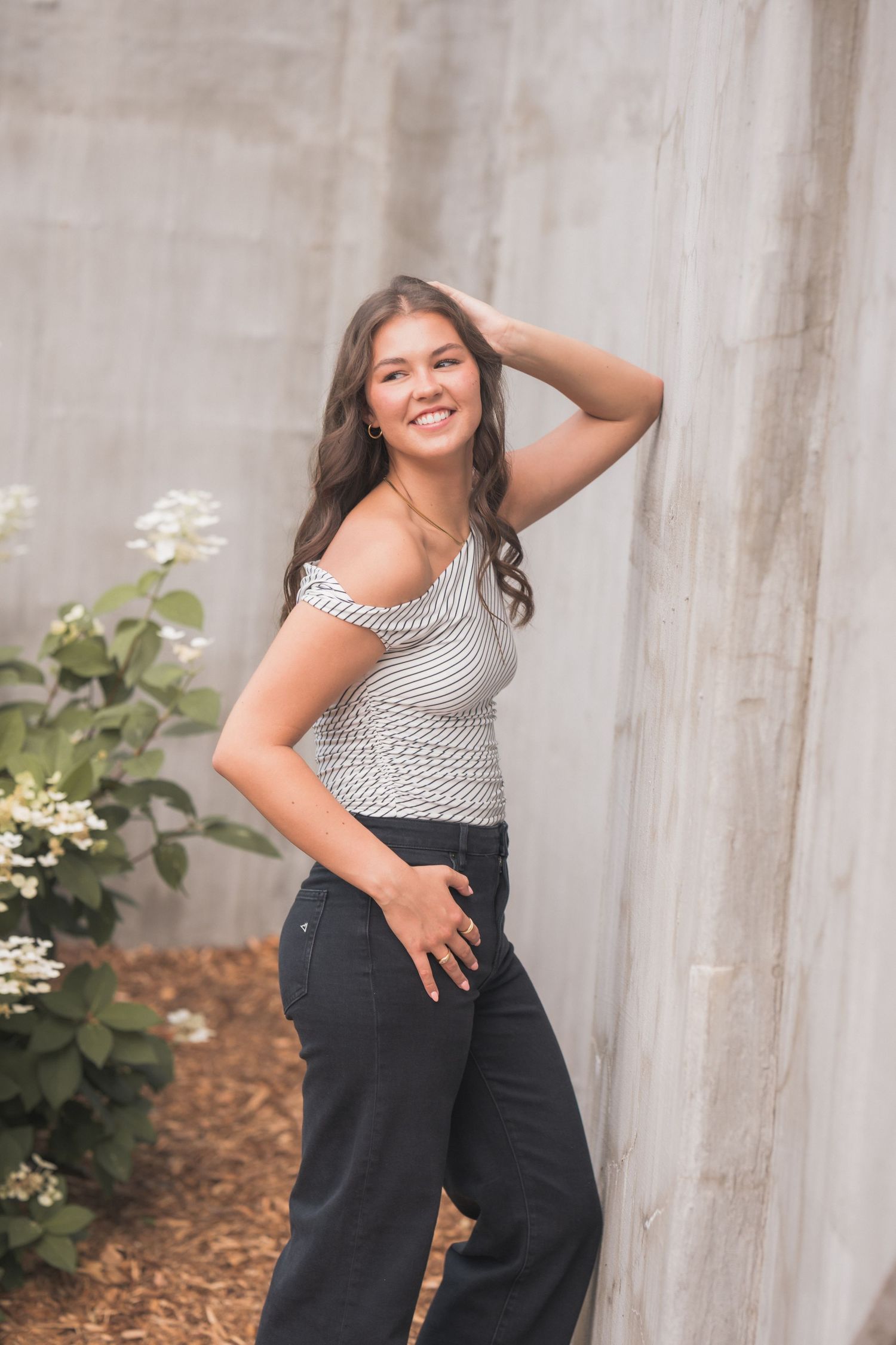Young woman in striped top and black jeans smiling, leaning against concrete wall with white flowers nearby.