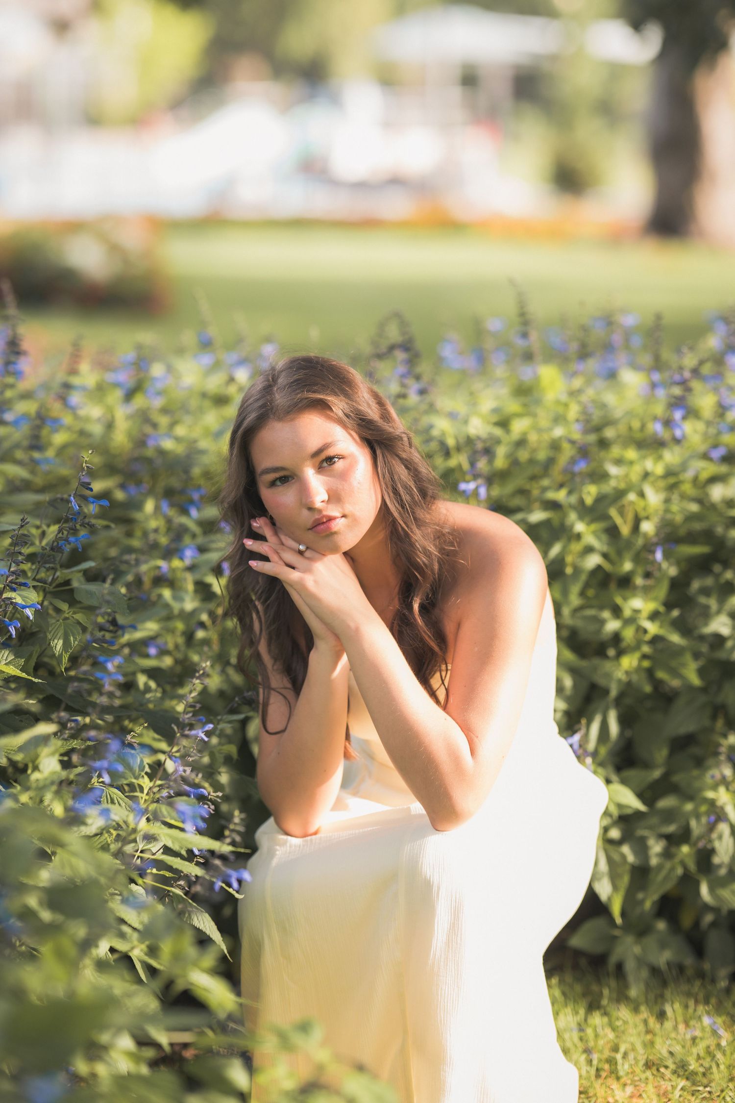 Young woman in white dress posing among blue flowers in a sunny garden park setting.