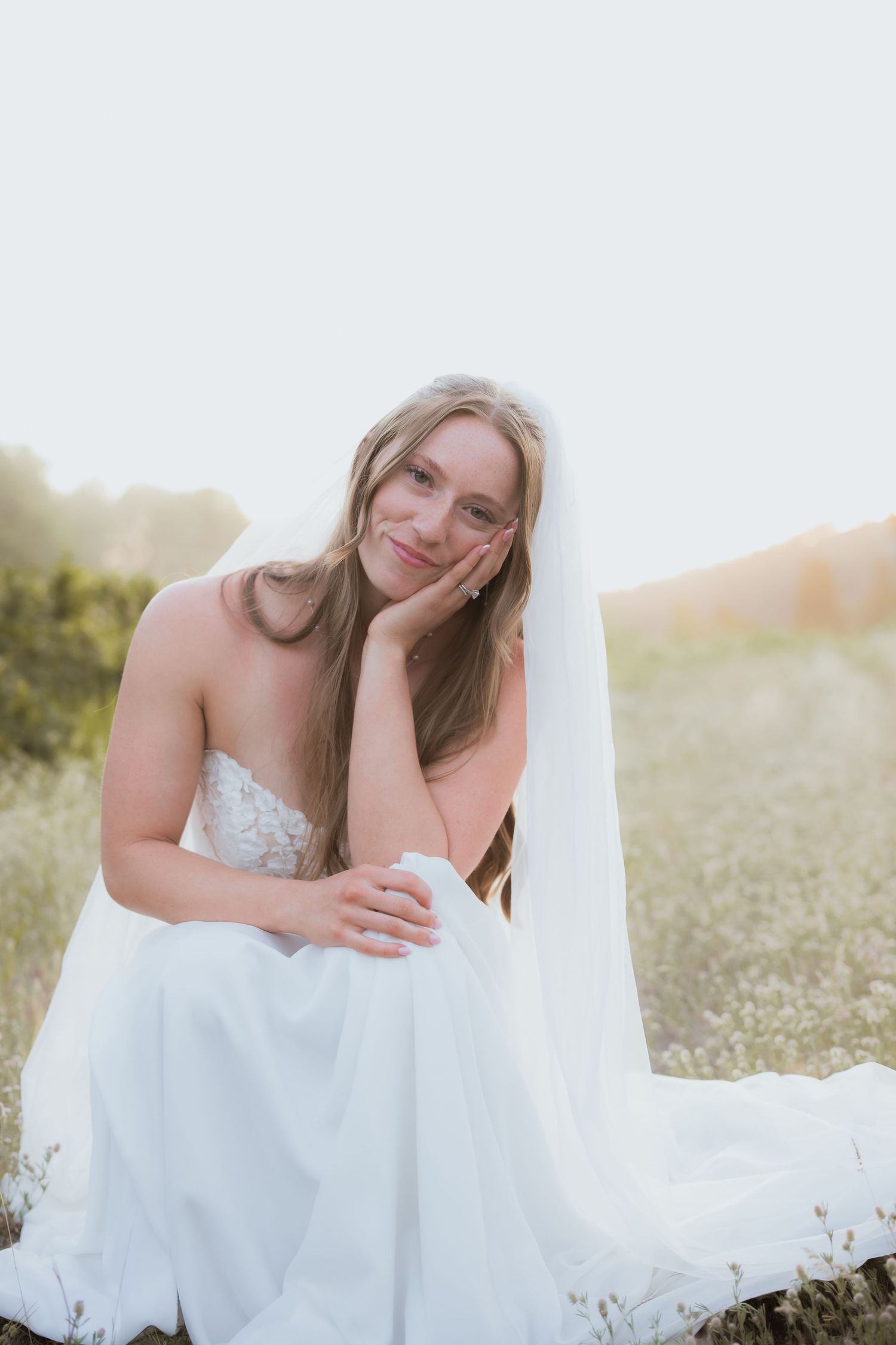 Blonde bride in a white strapless gown and veil sitting in a sunlit field, resting her cheek on her hand.