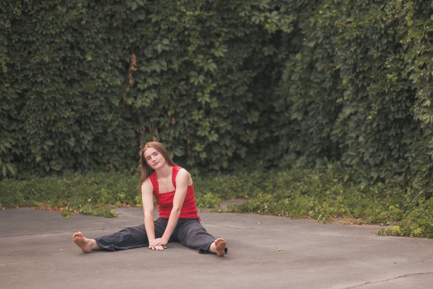 Young woman in red top and jeans doing splits stretch on pavement outdoors near green ivy wall.