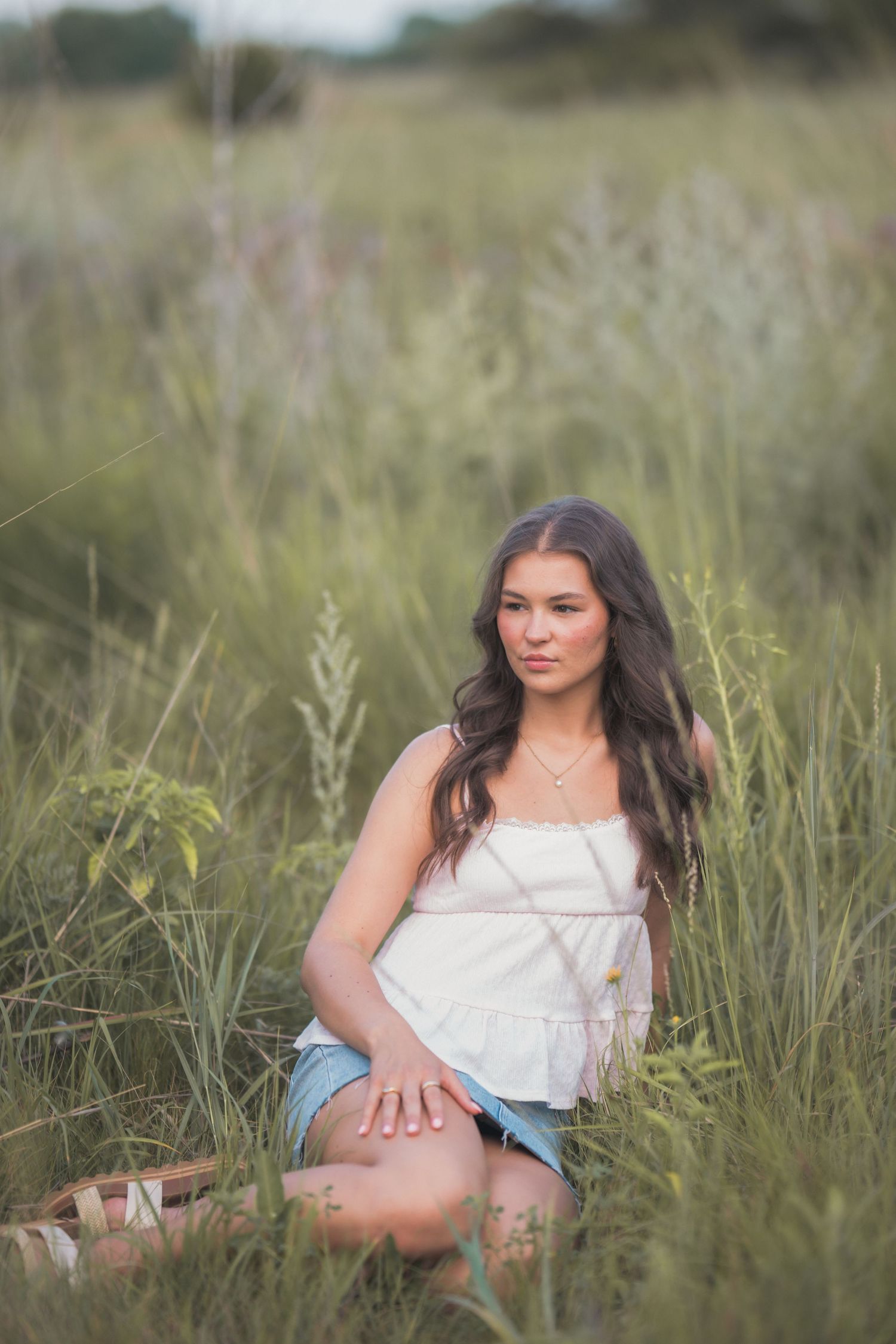 Young woman with long dark hair sits in tall green grass field wearing white top and denim shorts in golden hour light.