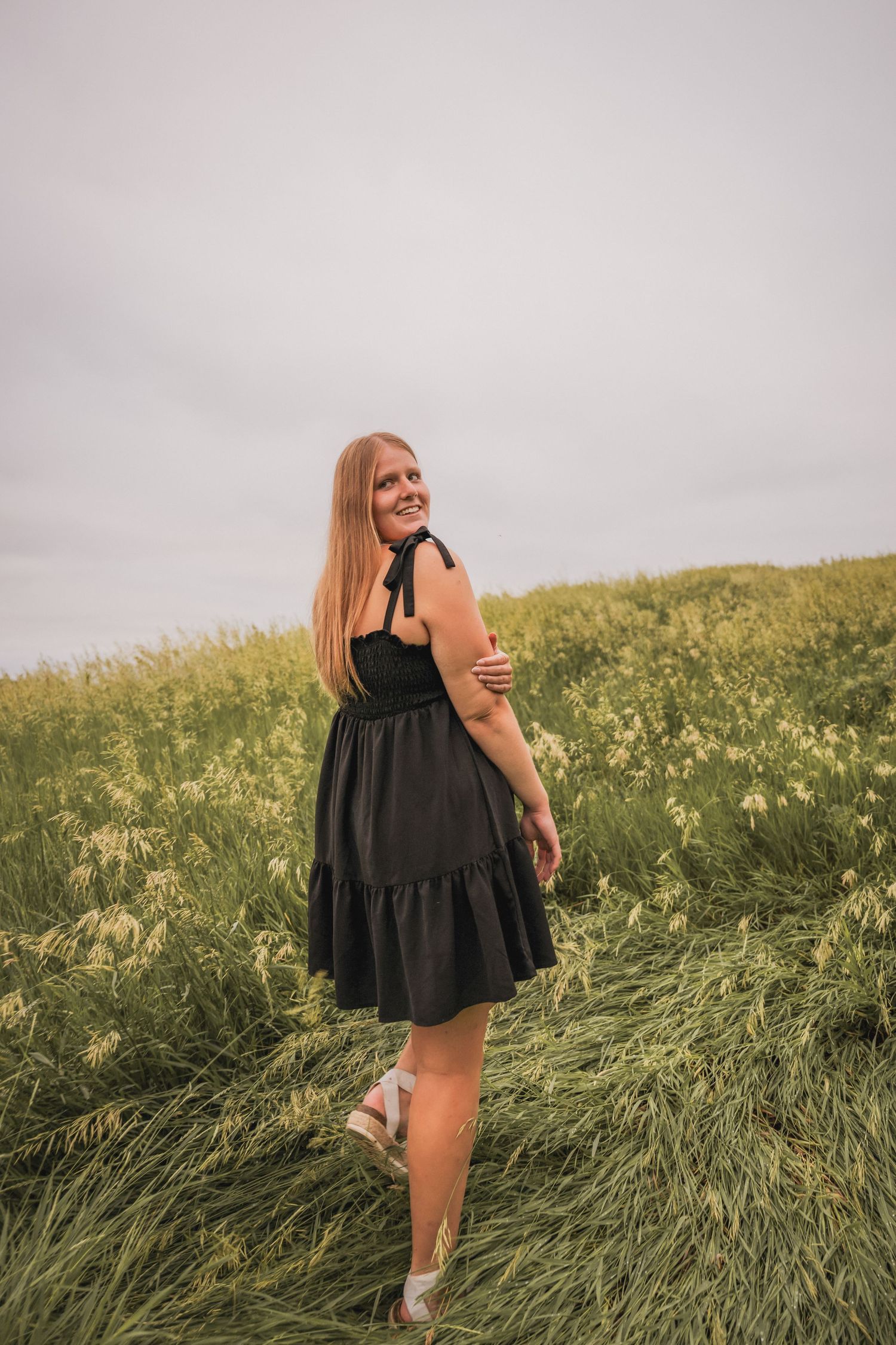Young woman in black sundress smiling over shoulder in green flower field.