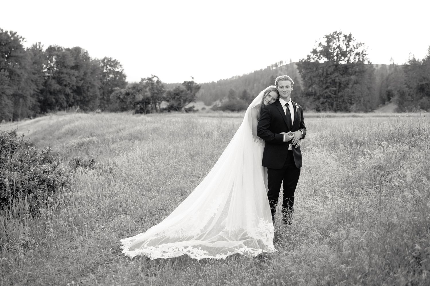 Black and white wedding portrait of bride and groom embracing in scenic green meadow.