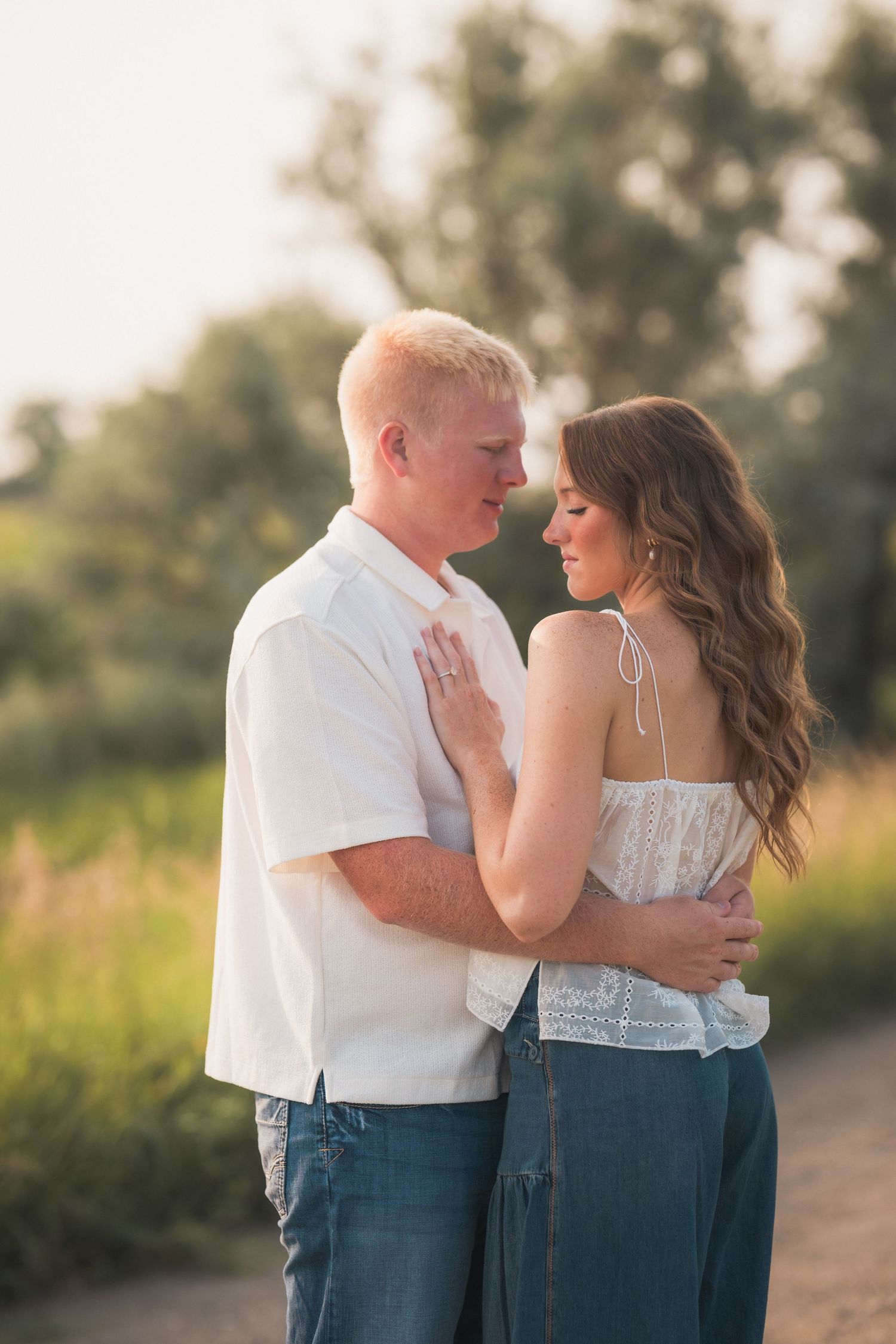 A couple in casual attire shares a romantic moment outdoors at golden hour, surrounded by lush greenery.