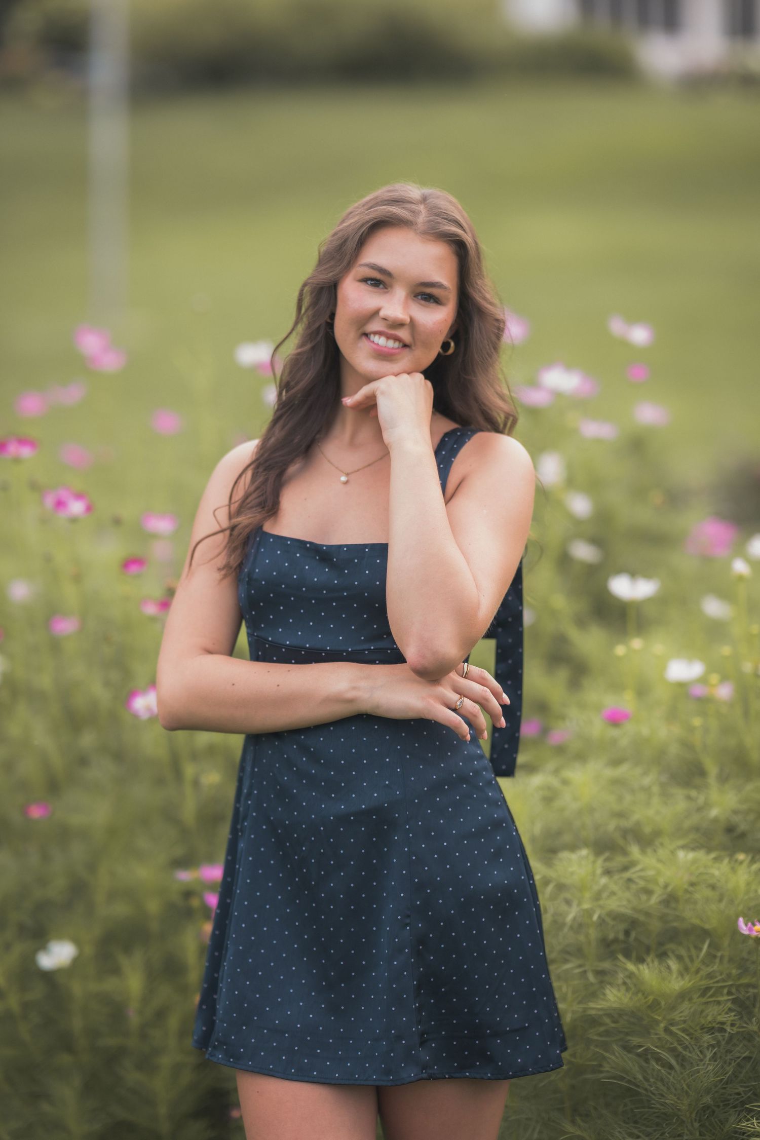 Young woman in navy polka dot dress smiling in a flower garden with pink cosmos blooms in background.
