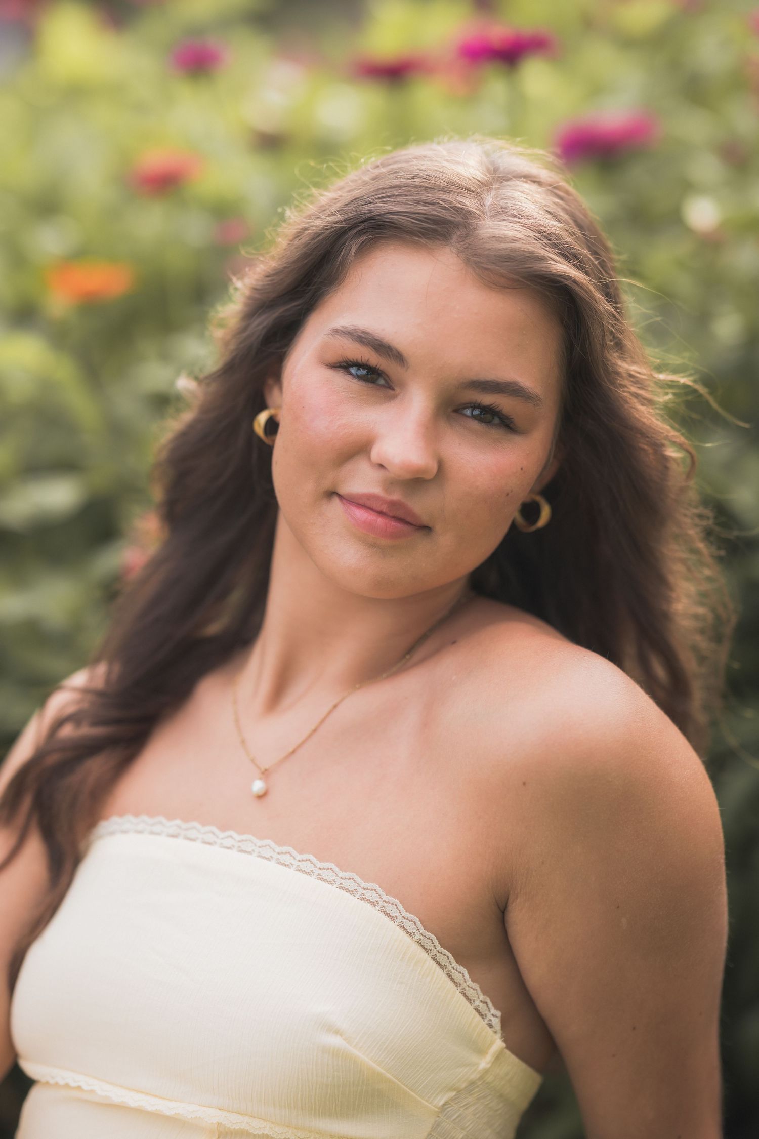 Young woman with long brown hair wearing a yellow strapless top and pearl necklace posing in a flower garden.