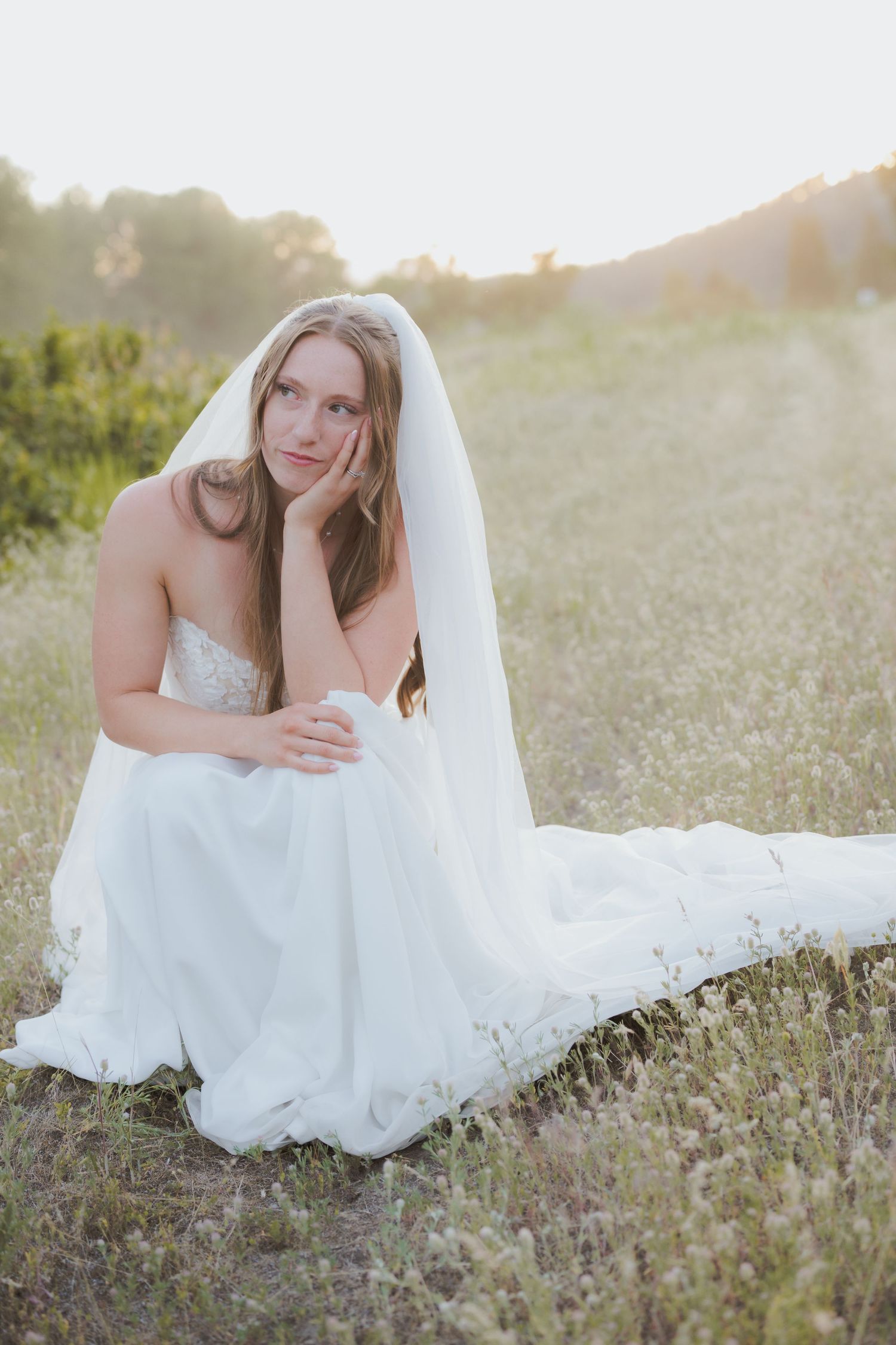 Bride in white gown and veil sitting in a sunlit meadow, resting her chin on her hand.