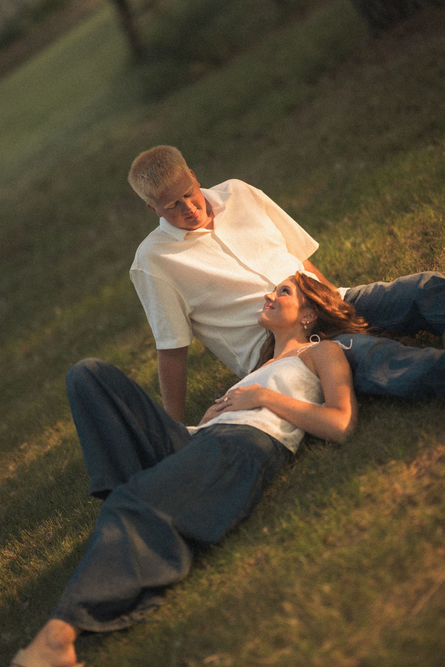 Couple relaxing on green grass at sunset, woman lying down smiling up at man seated beside her outdoors.