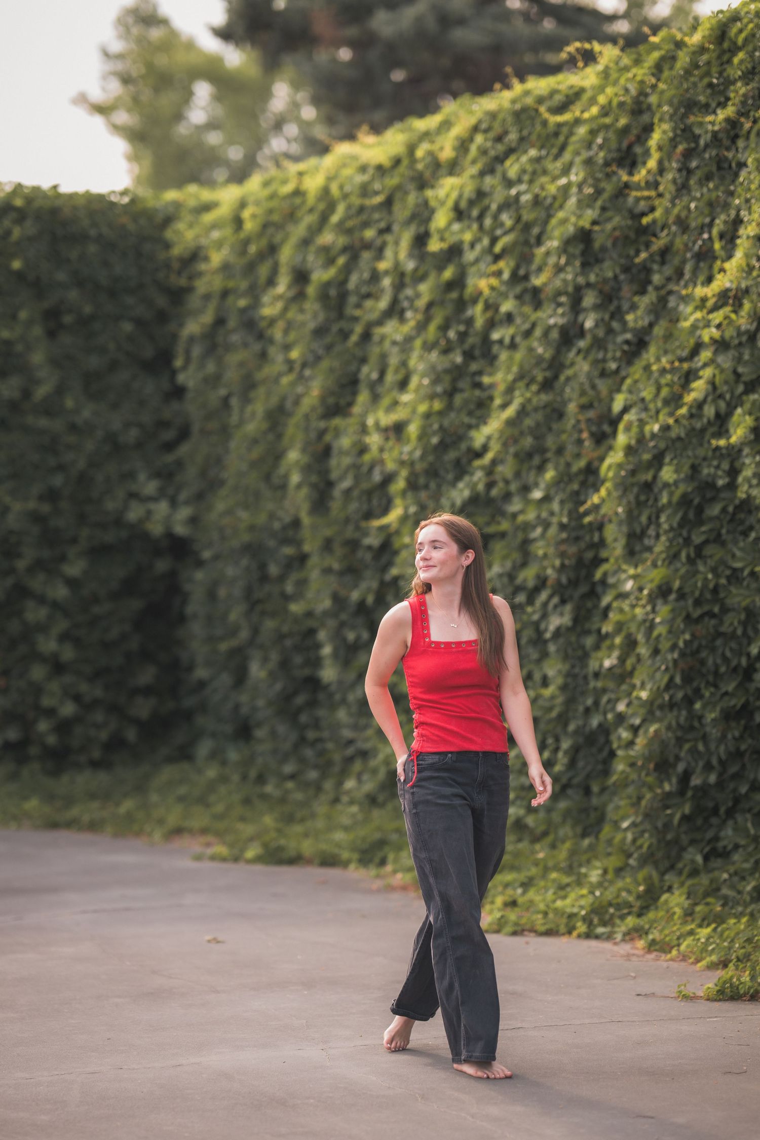 A smiling young woman in a red tank top and dark jeans walks barefoot along a path beside a lush green ivy-covered wall.