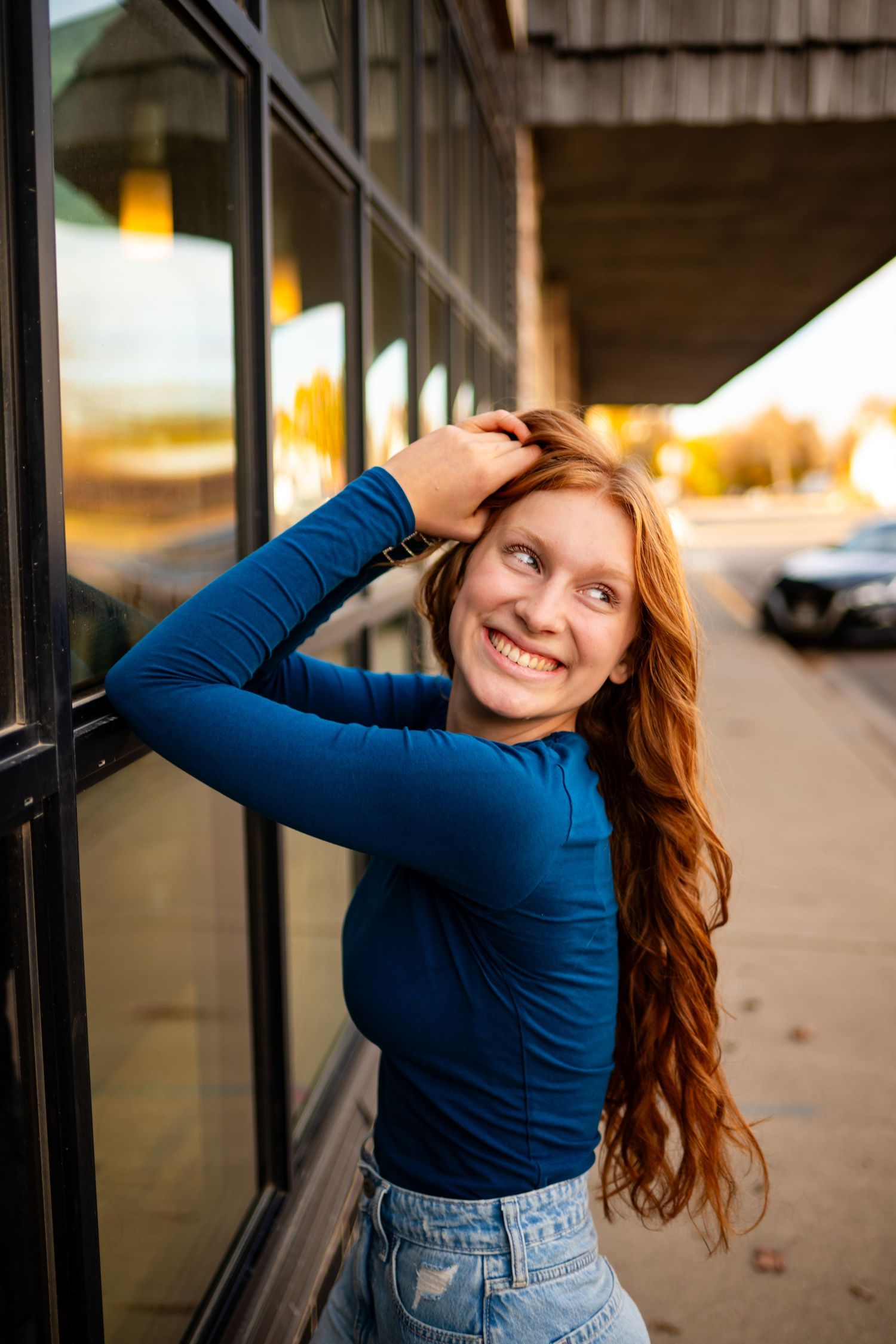 Smiling redhead teen girl in blue top leaning against storefront window on sidewalk.