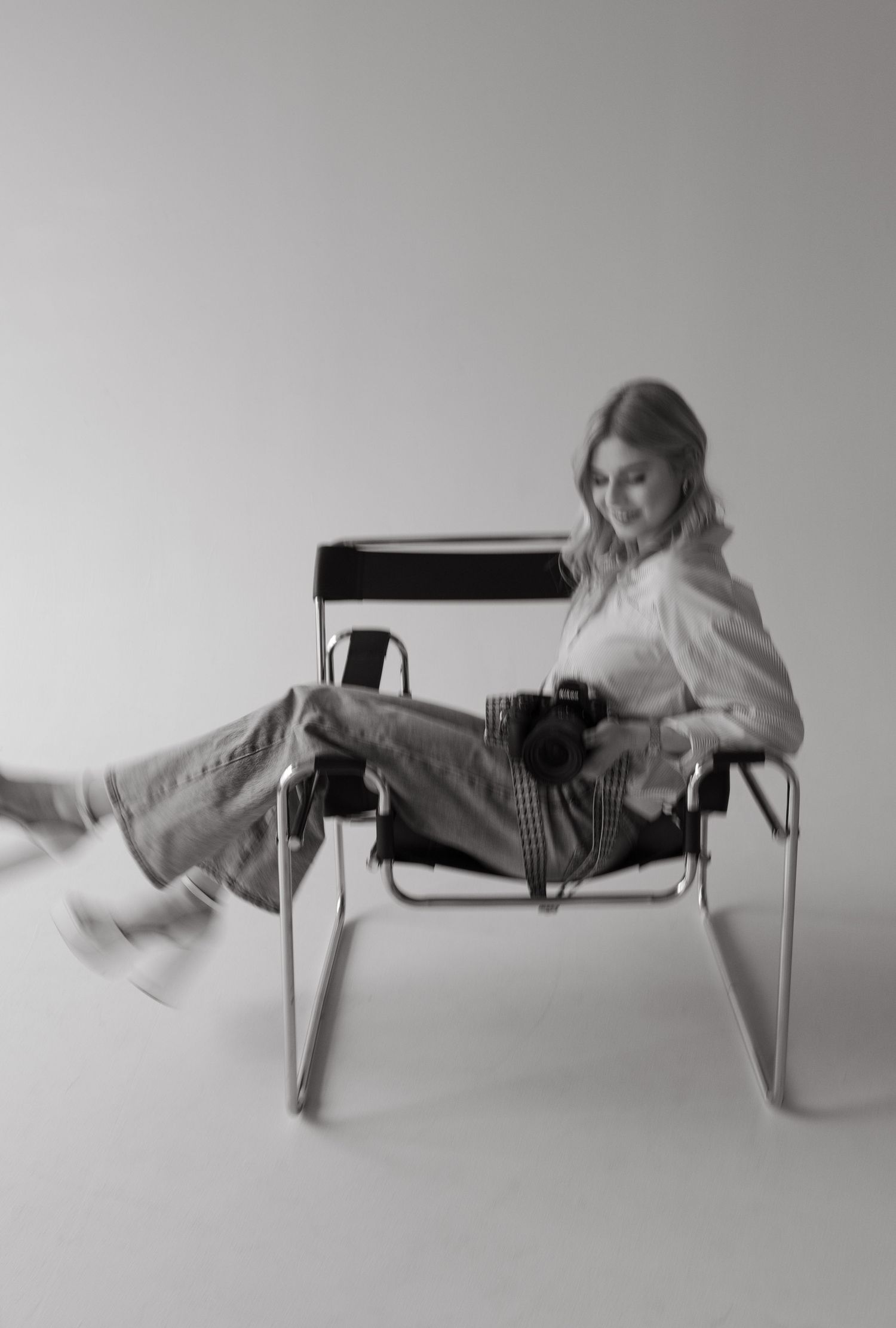 Black and white photo of Joclyn Rae sitting in a Wassily chair holding a camera, wearing jeans and a white shirt.