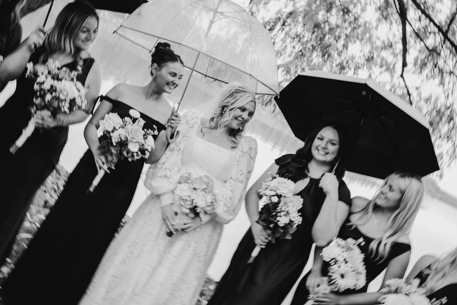 Black and white photo of bride with bridesmaids holding umbrellas in the rain on wedding day.