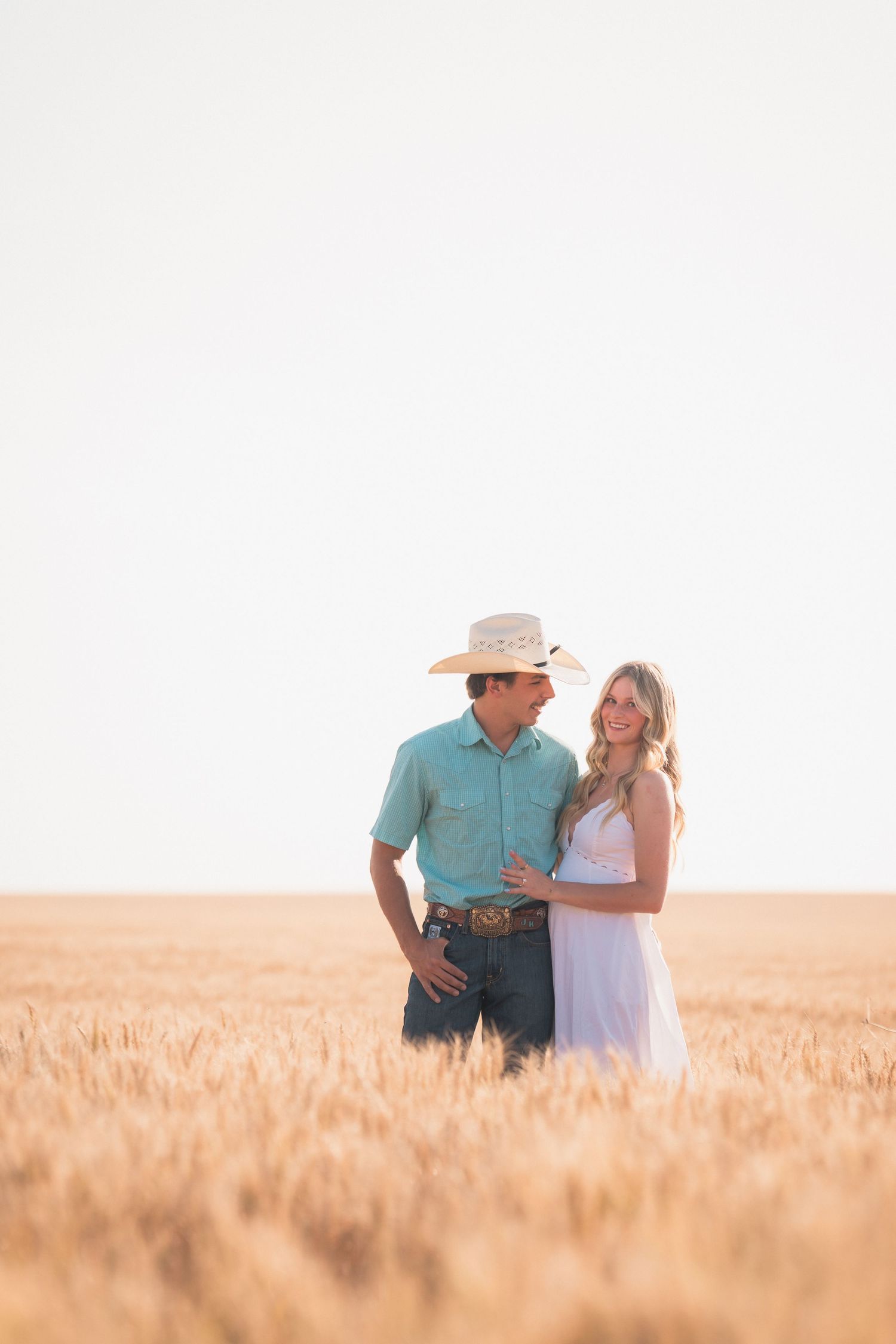 Couple embracing in golden wheat field, man in cowboy hat and teal shirt, woman in white dress, sunny day.