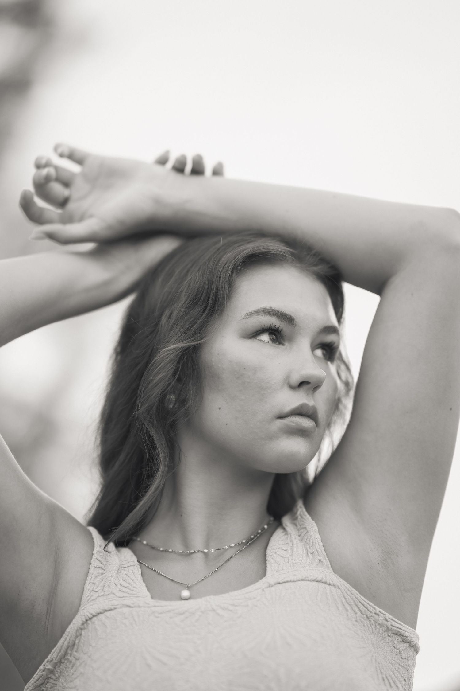Black and white portrait of a young woman with freckles, arms raised above head, wearing a delicate necklace.