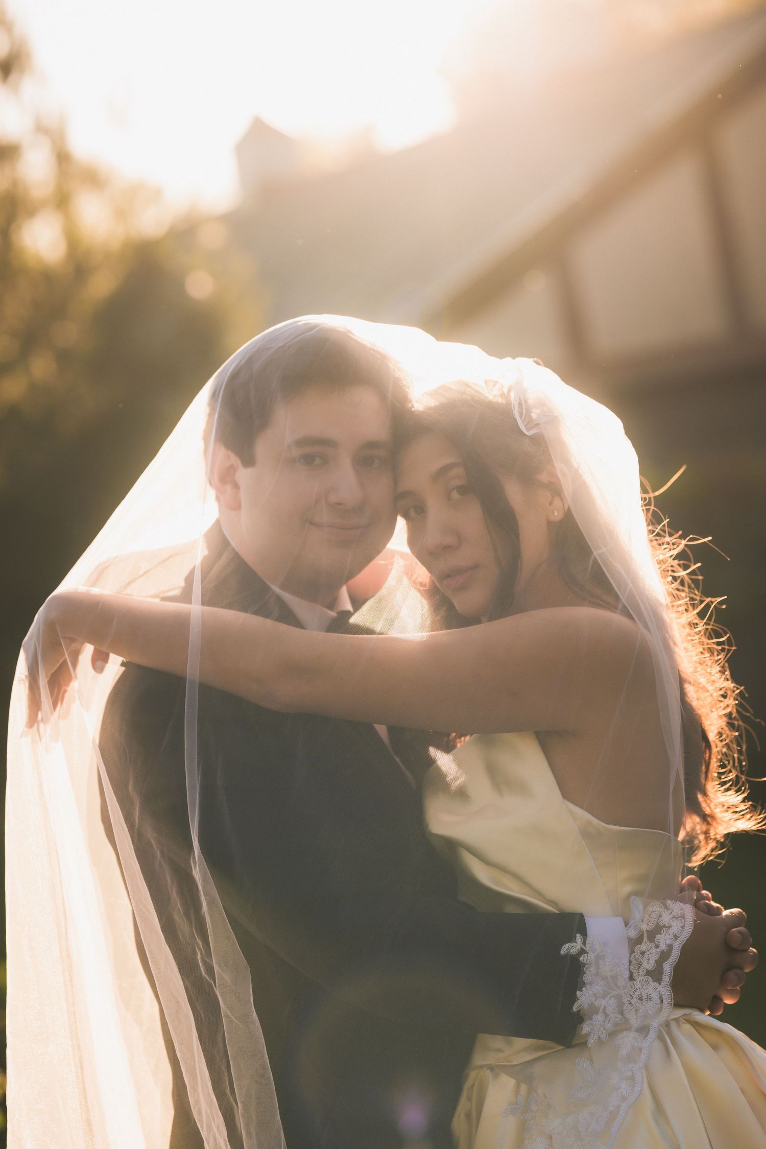 Bride and groom embrace under a sheer wedding veil in warm golden sunset light outside a timber-framed building.