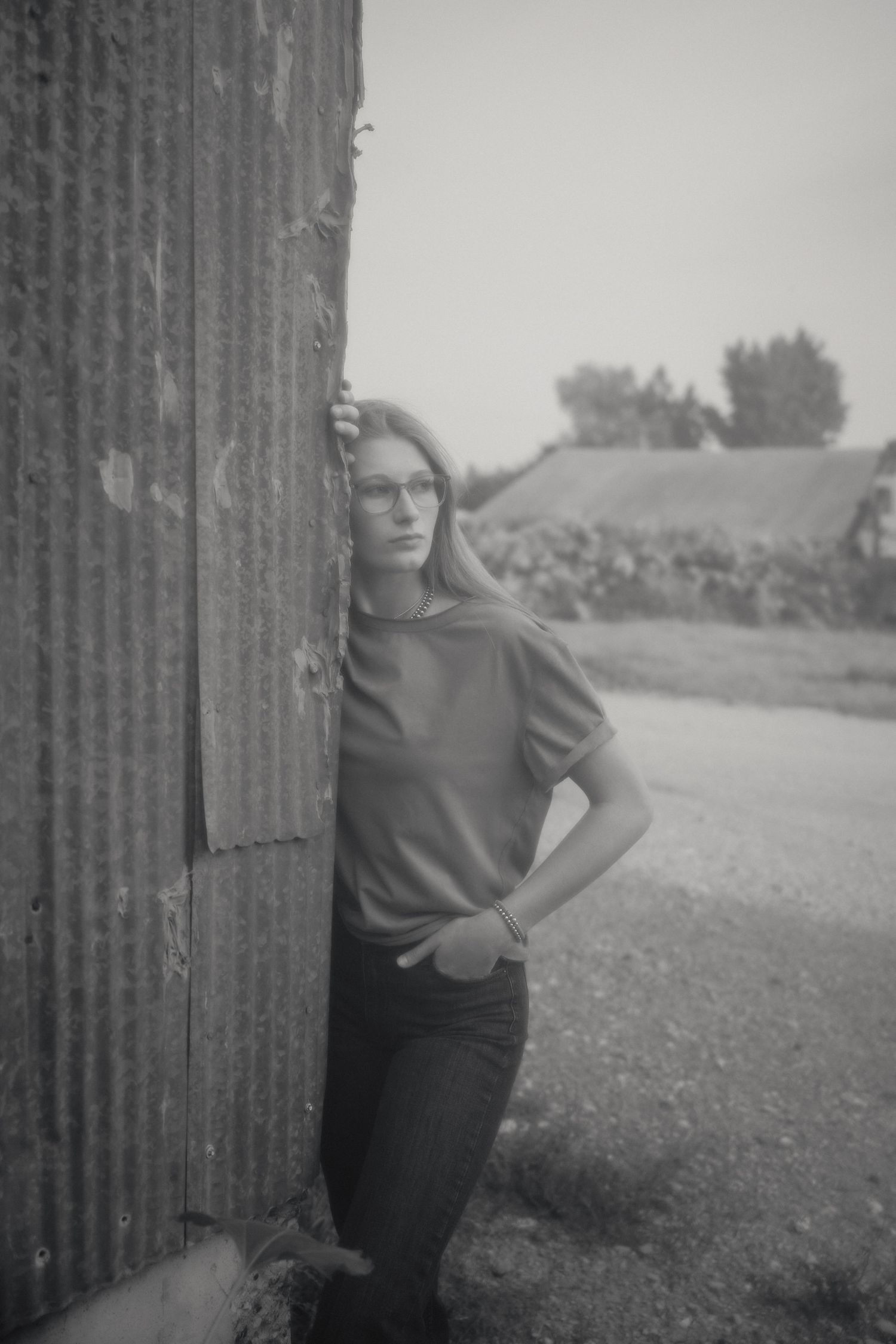 Blonde woman with glasses leaning against rustic corrugated wall in black and white portrait.