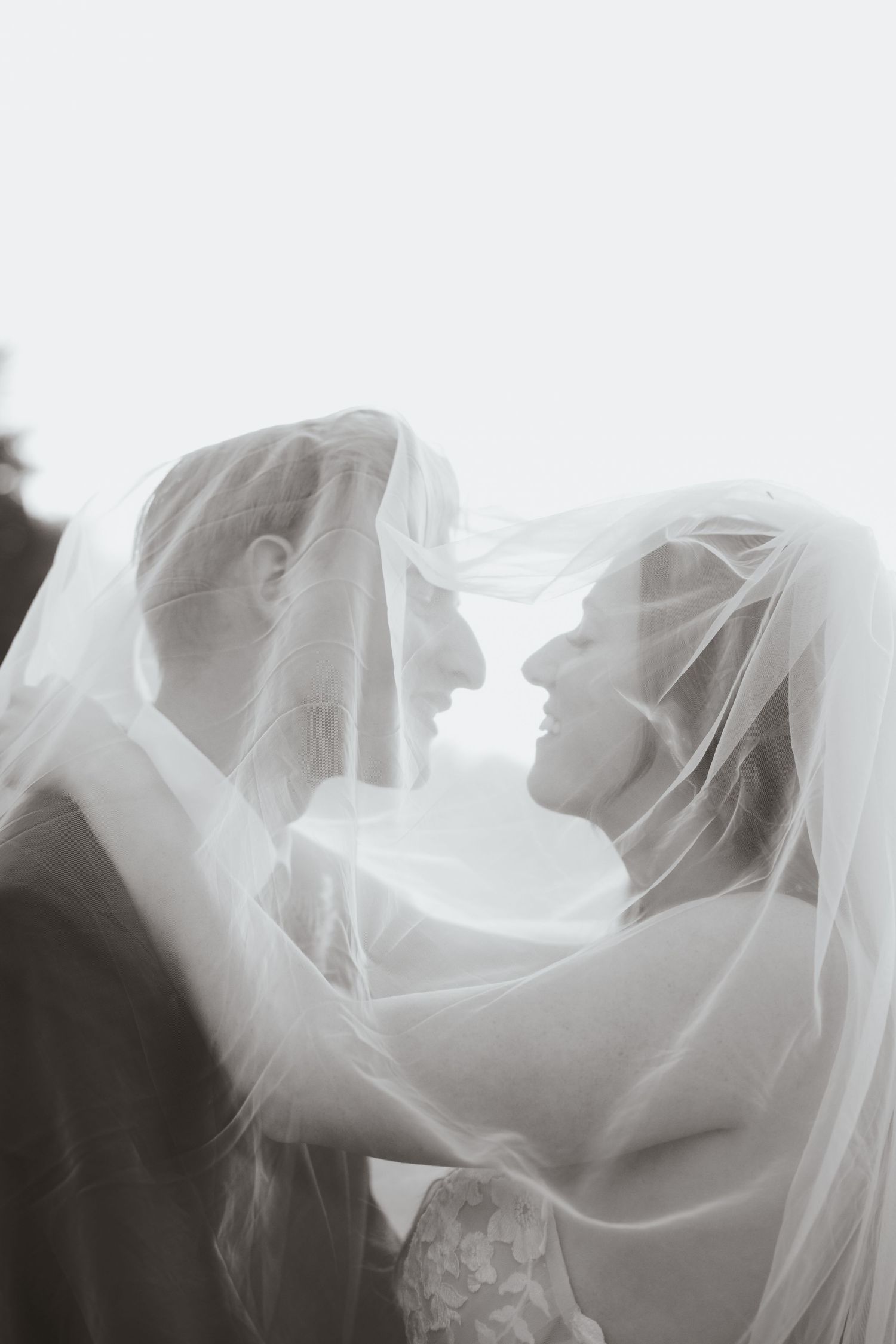 Black and white photo of bride and groom sharing romantic moment under flowing wedding veil.