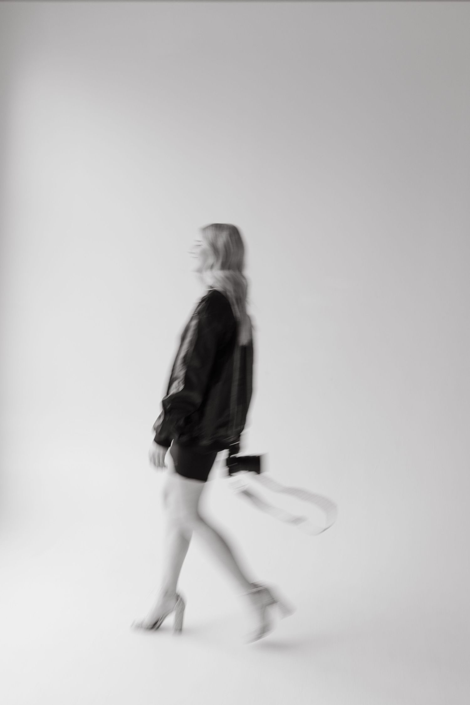 Black and white motion blur photo of woman in black jacket and heels walking in white studio.