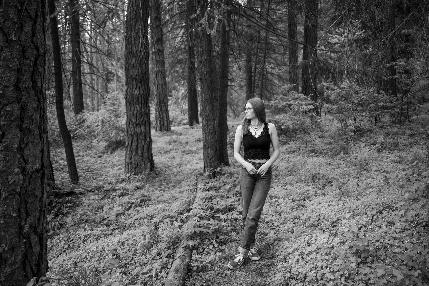 Woman in black top standing among tall pine trees in black and white forest photo.