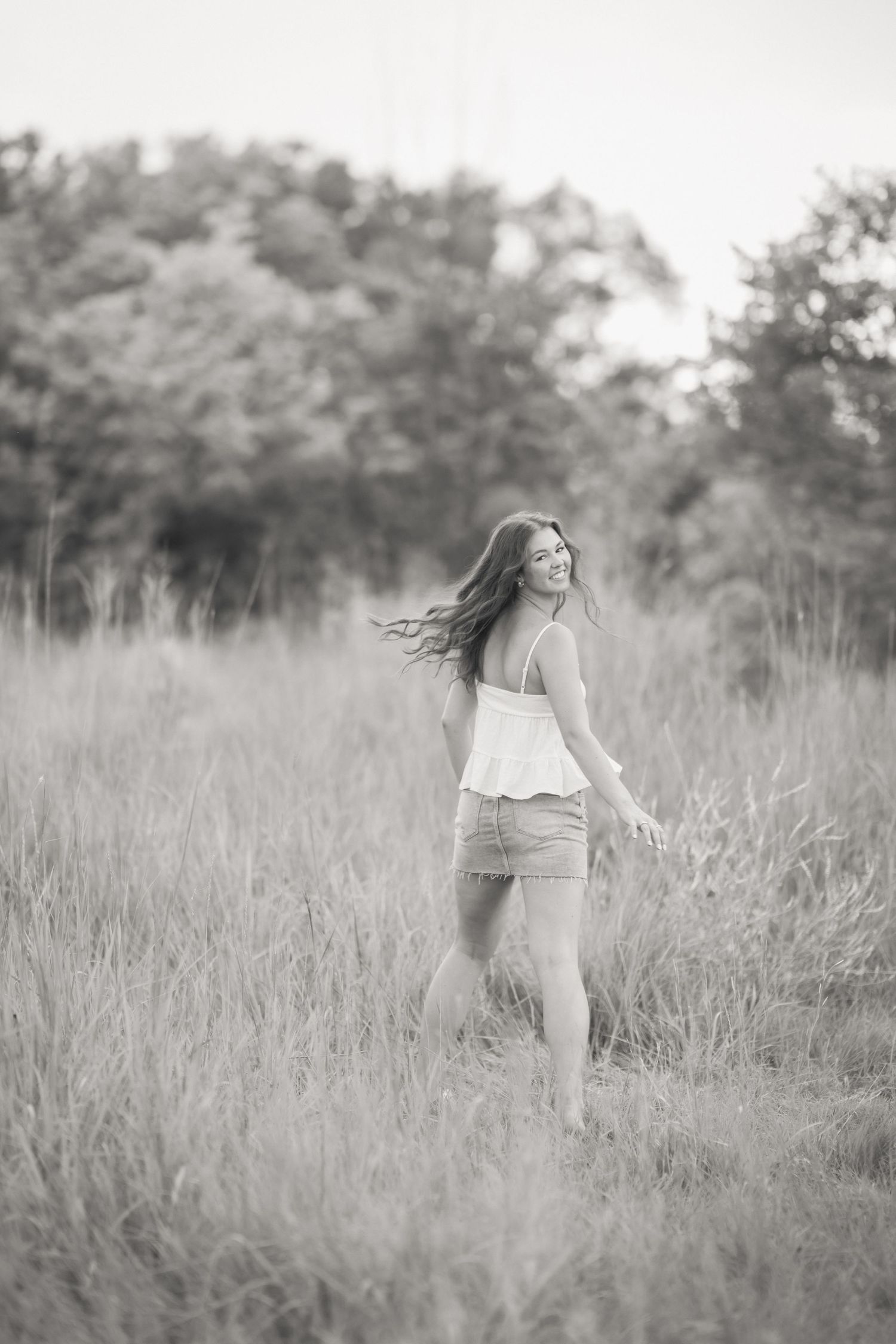 Black and white photo of a young woman in a white top and shorts standing in a tall grass field with trees behind her.