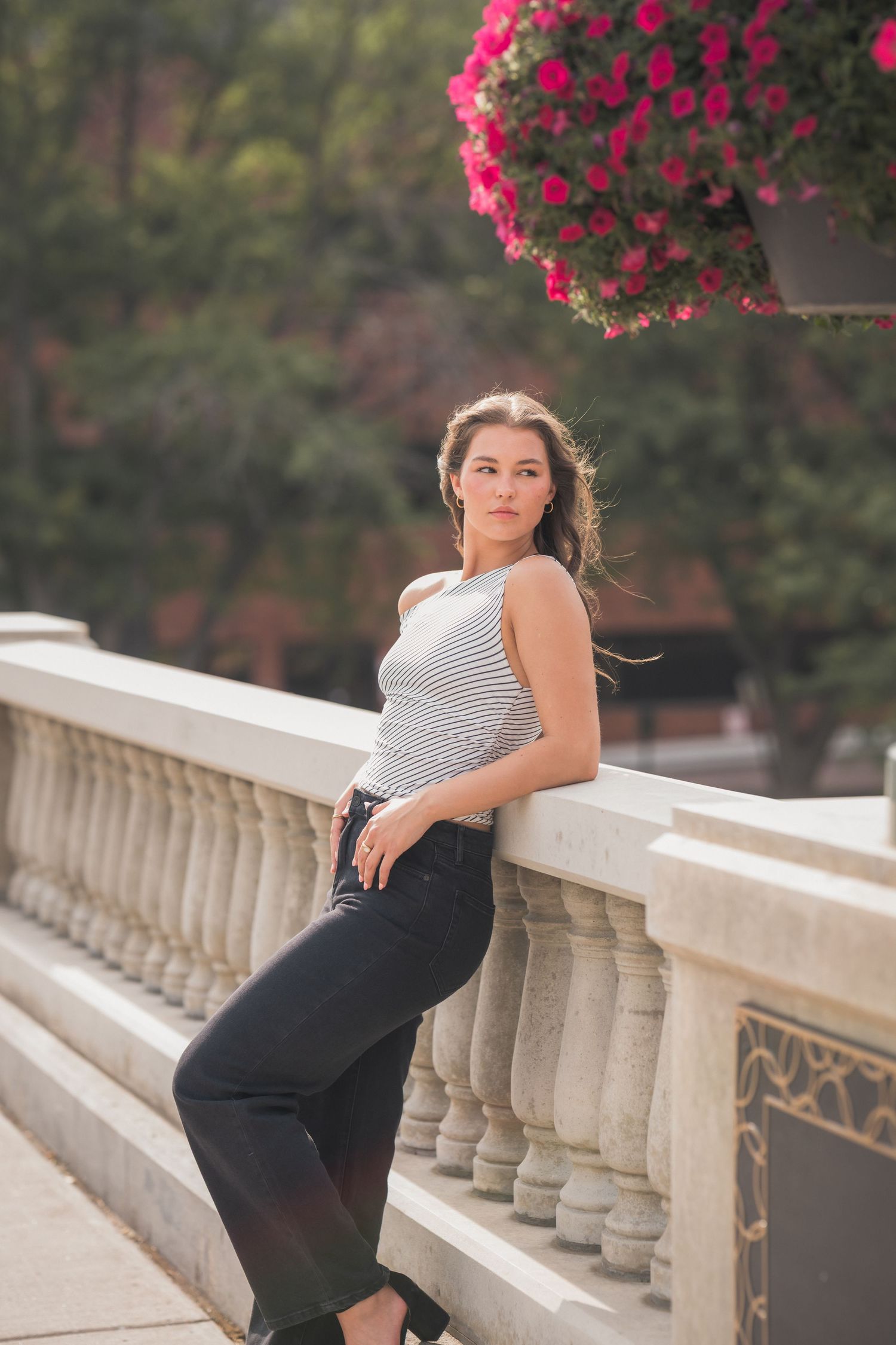 Young woman in striped top and black jeans leaning on stone balustrade with pink flowers in urban setting.