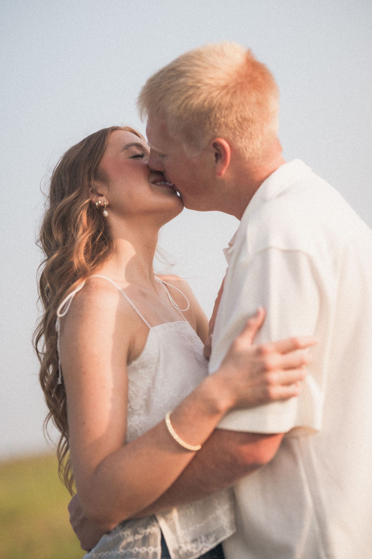 A couple sharing a romantic kiss outdoors, woman in white dress with long wavy hair, man in white shirt.