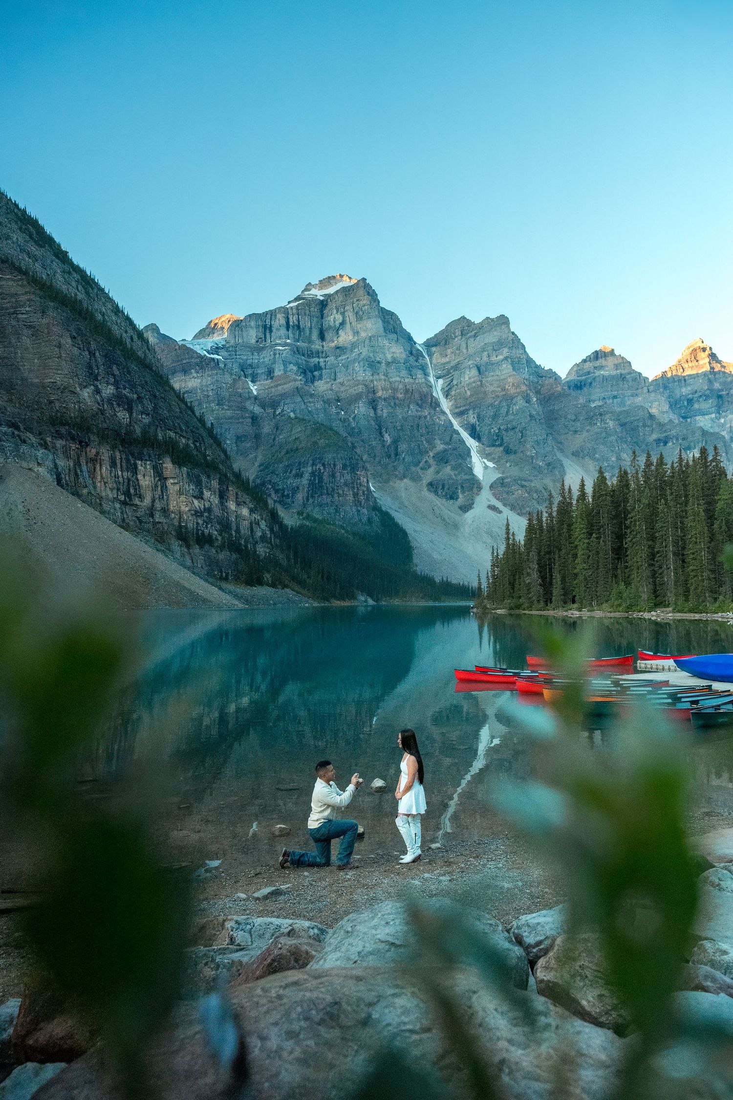 Man proposing at sunrise beside Moraine Lake with red canoes and golden mountain reflections in water.
