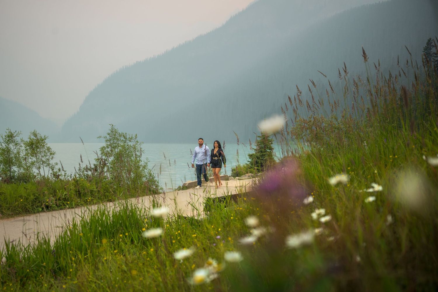 Couple walking hand in hand on a scenic lakeside path surrounded by wildflowers and mountain views.