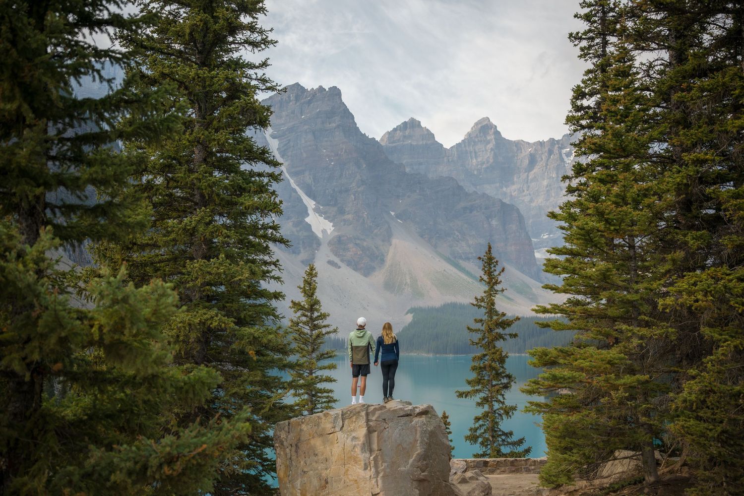 Two hikers stand on a rocky ledge overlooking turquoise Moraine Lake with mountain peaks and pine trees in Banff.