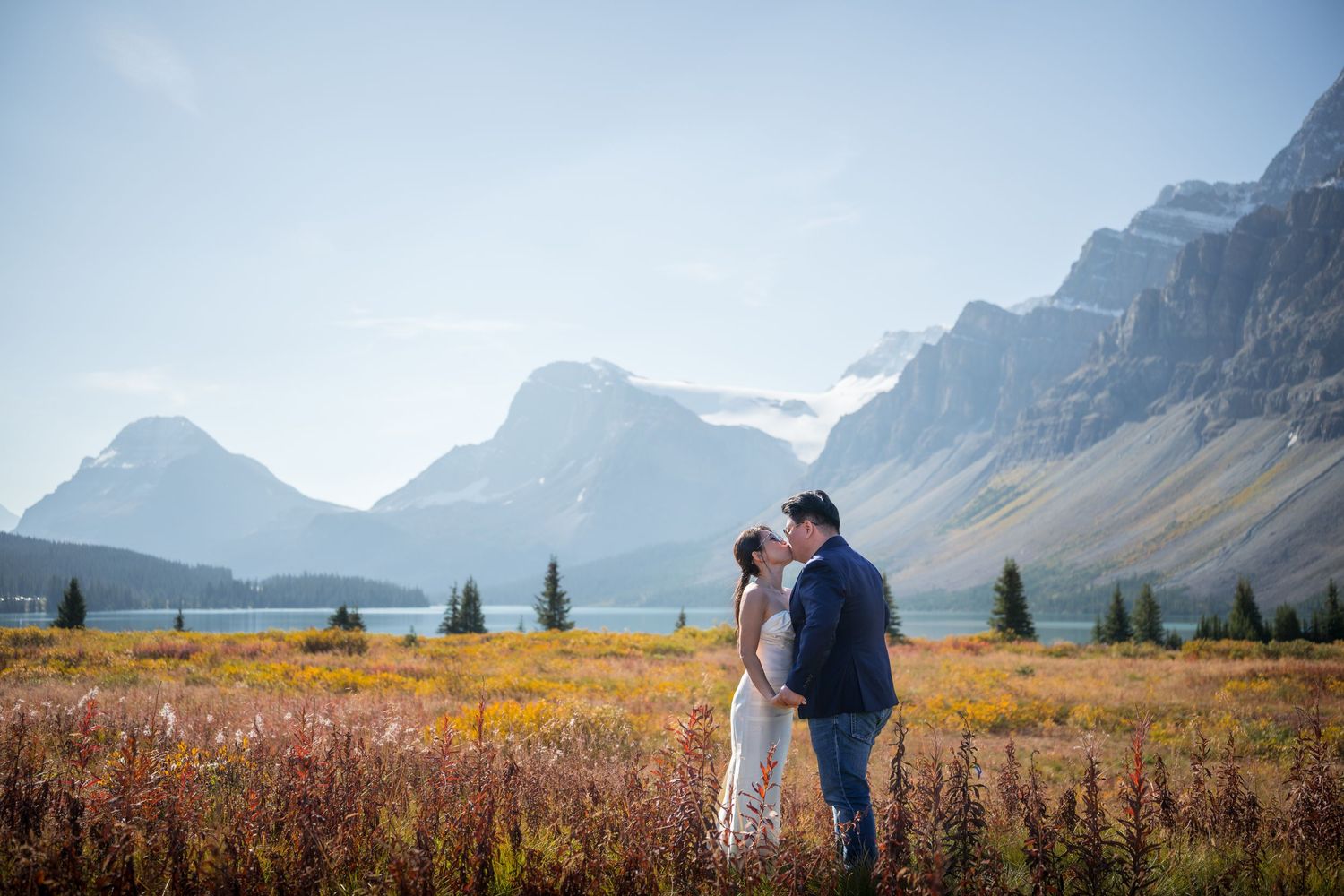 Couple sharing a romantic kiss in a colorful mountain meadow with a serene lake and majestic peaks in the background.