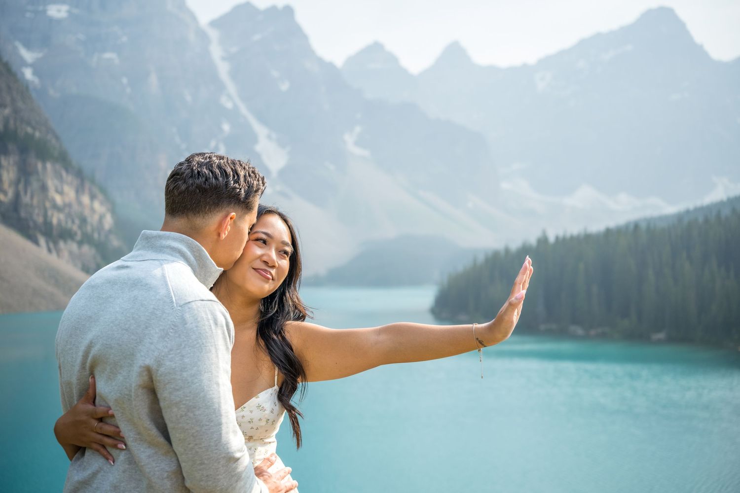 Couple embracing at turquoise Moraine Lake with Rocky Mountain peaks in background, bride waving at camera.