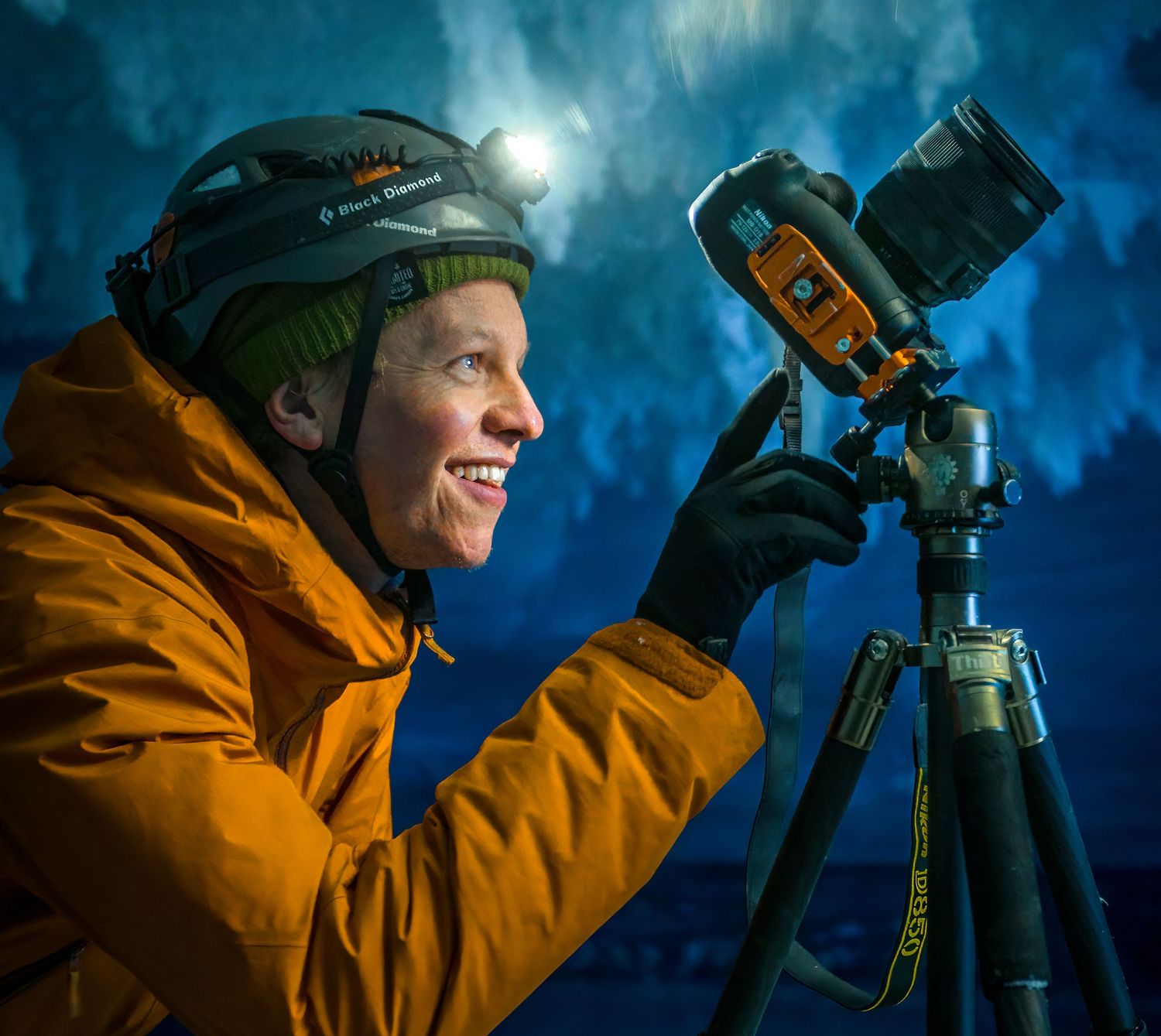 Smiling photographer in orange jacket using camera on tripod in a blue ice cave with headlamp.