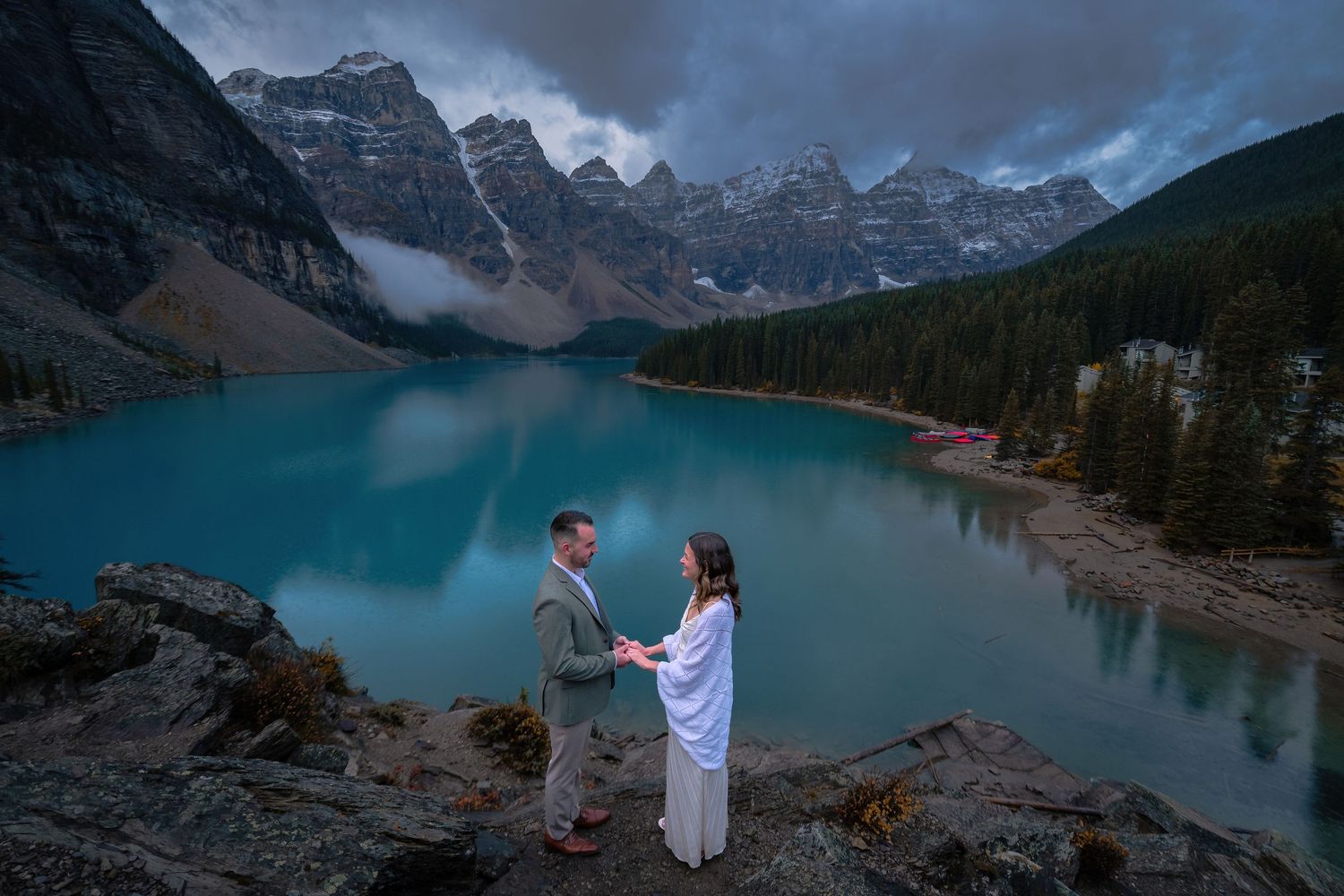 Couple holding hands during elopement ceremony at Moraine Lake with dramatic mountain backdrop at dusk.