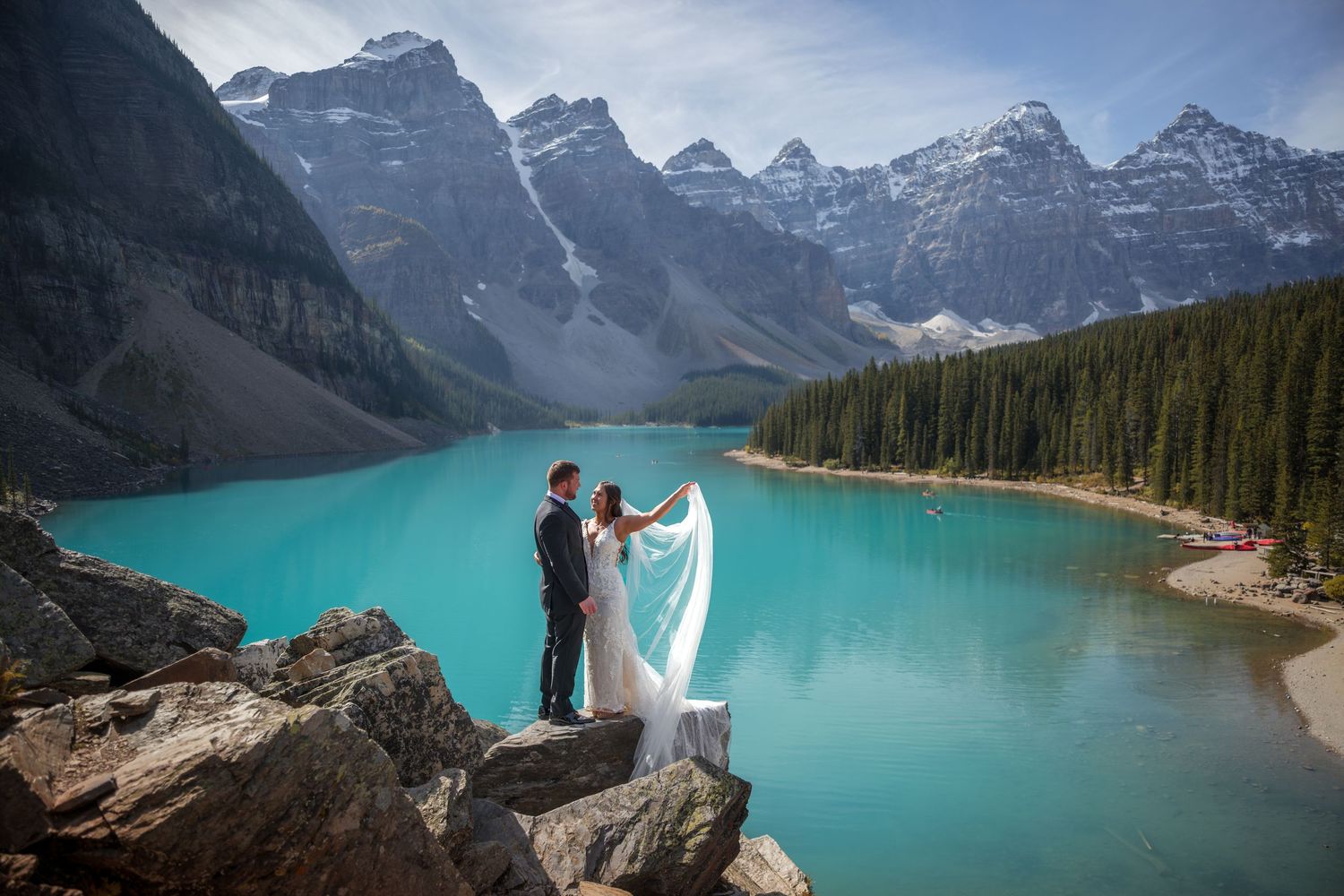 Bride and groom standing on rocky shore of turquoise Moraine Lake with snow-capped Canadian Rockies backdrop.