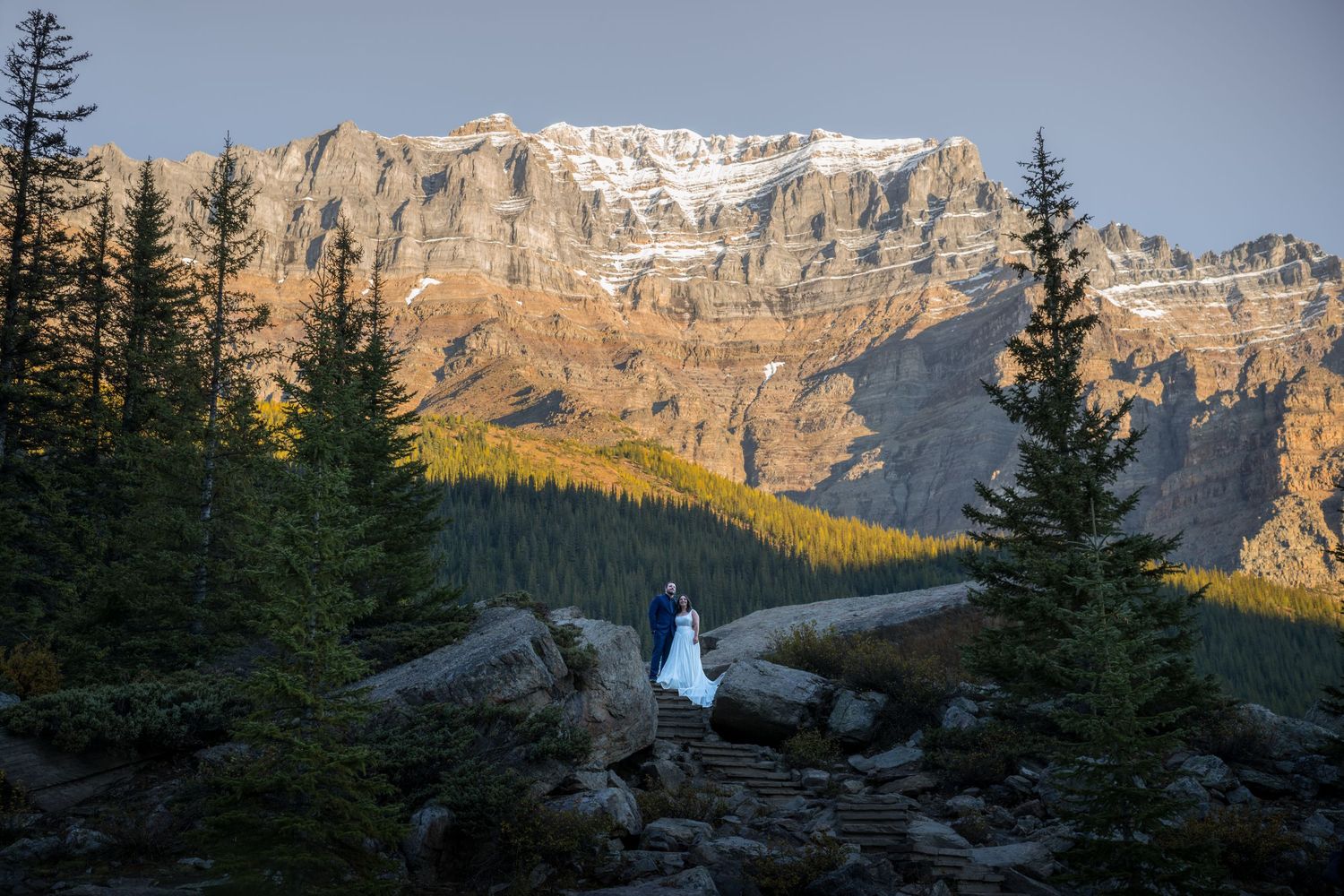 Bride and groom stand among pine trees with stunning snow-capped Rocky Mountain peaks in the background at a mountain wedding.