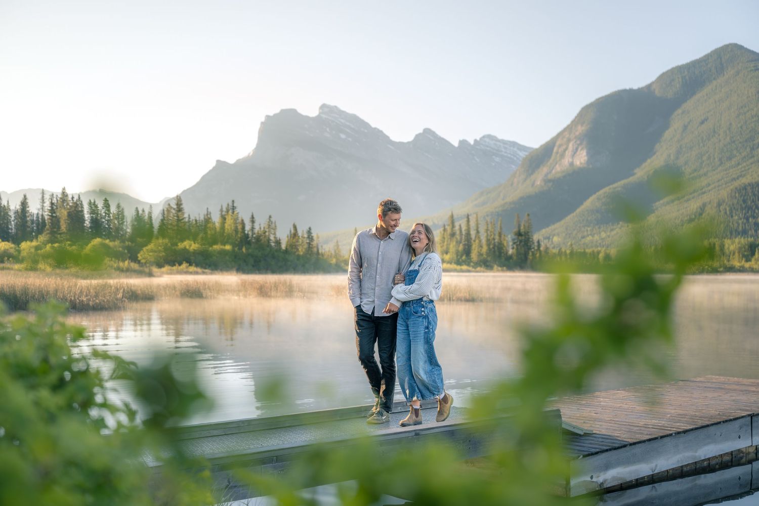 Couple walking hand in hand on a wooden dock with a serene mountain lake and lush green trees in the background.