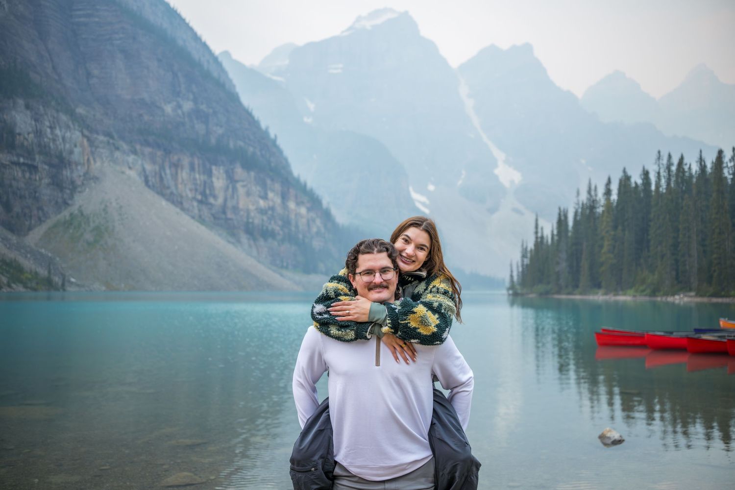 Couple smiling at Moraine Lake in Banff, Canada, with turquoise water, mountains, and a red canoe in the background.