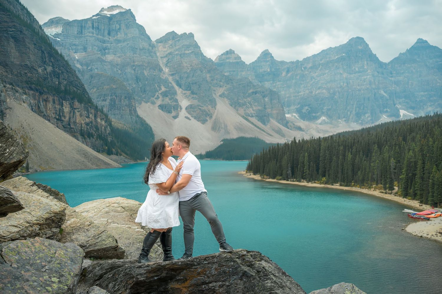 Couple kissing on rocks at Moraine Lake with stunning turquoise water and mountain peaks behind them.
