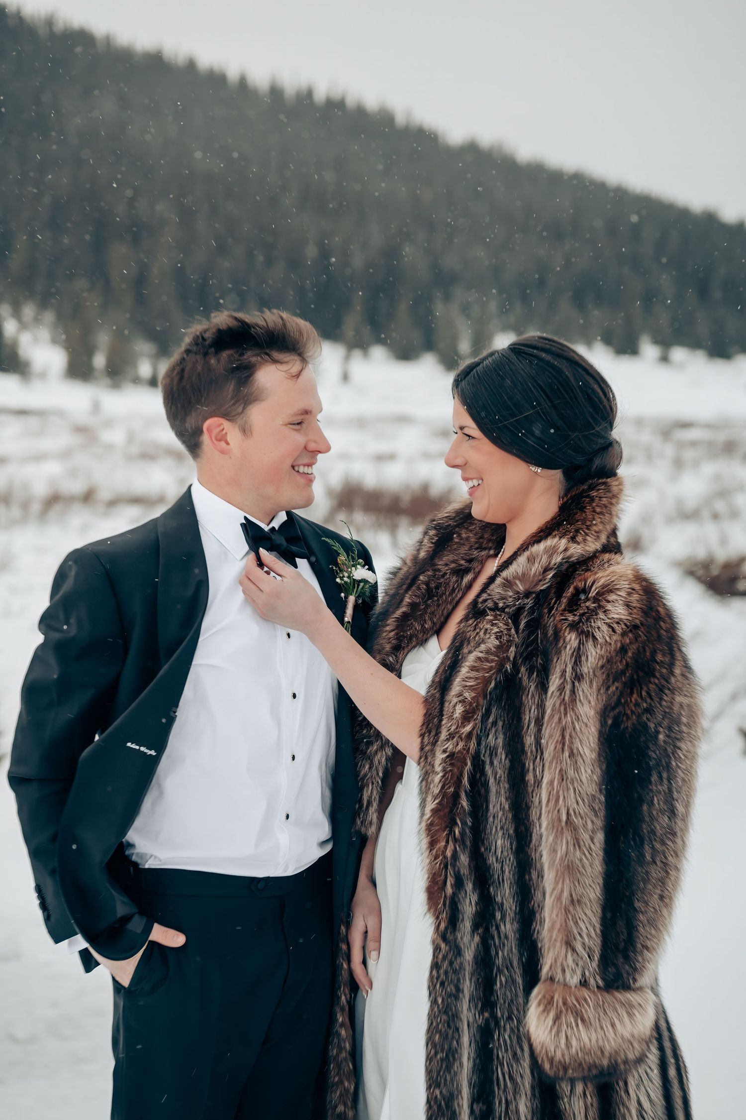 Bride in fur coat adjusts groom's bow tie during snowy winter wedding portrait outdoors.