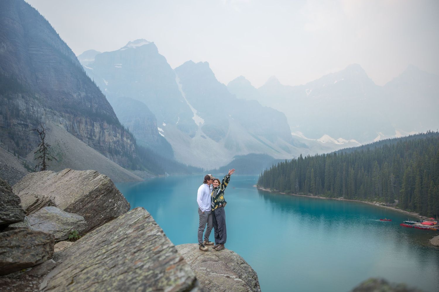Couple embracing on rocky ledge overlooking turquoise Moraine Lake with misty mountain peaks in Banff National Park.