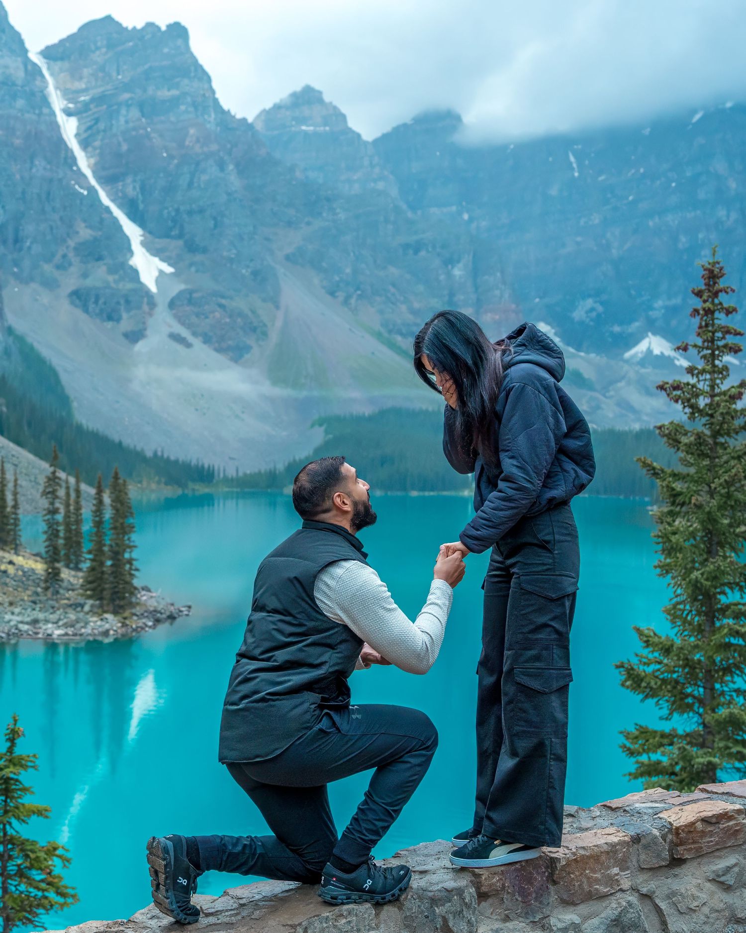 Man proposing to woman on rocky ledge at Moraine Lake with vivid turquoise water and pine trees.