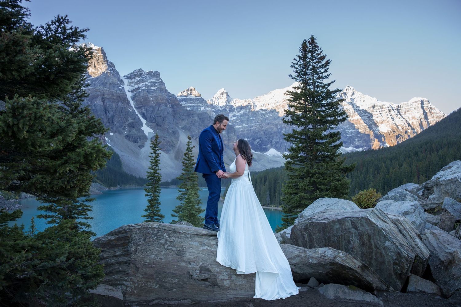 Couple in wedding attire holding hands at Moraine Lake, Banff, with turquoise water and Rocky Mountains backdrop.