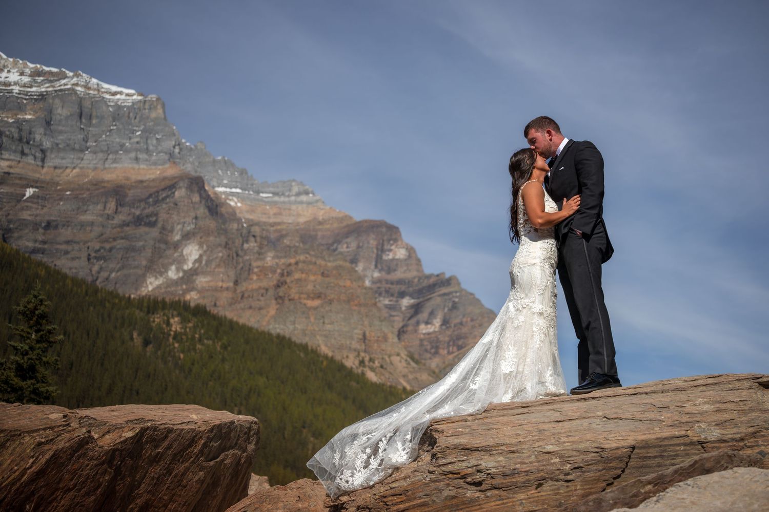 Bride and groom kissing on rocky mountain overlook with stunning peaks and blue sky backdrop.
