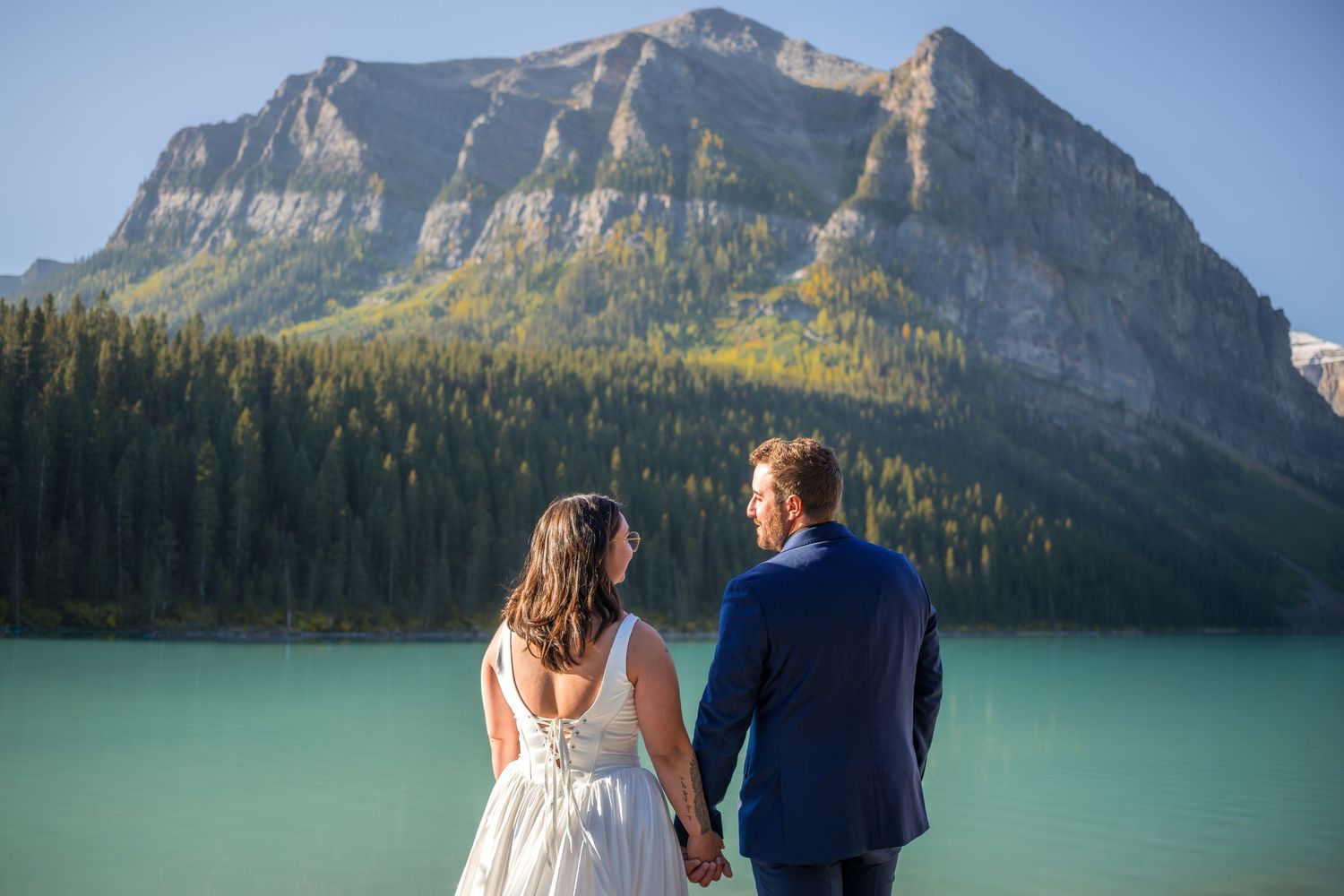 Bride and groom holding hands facing turquoise Lake Louise with majestic Rocky Mountain backdrop on their wedding day.