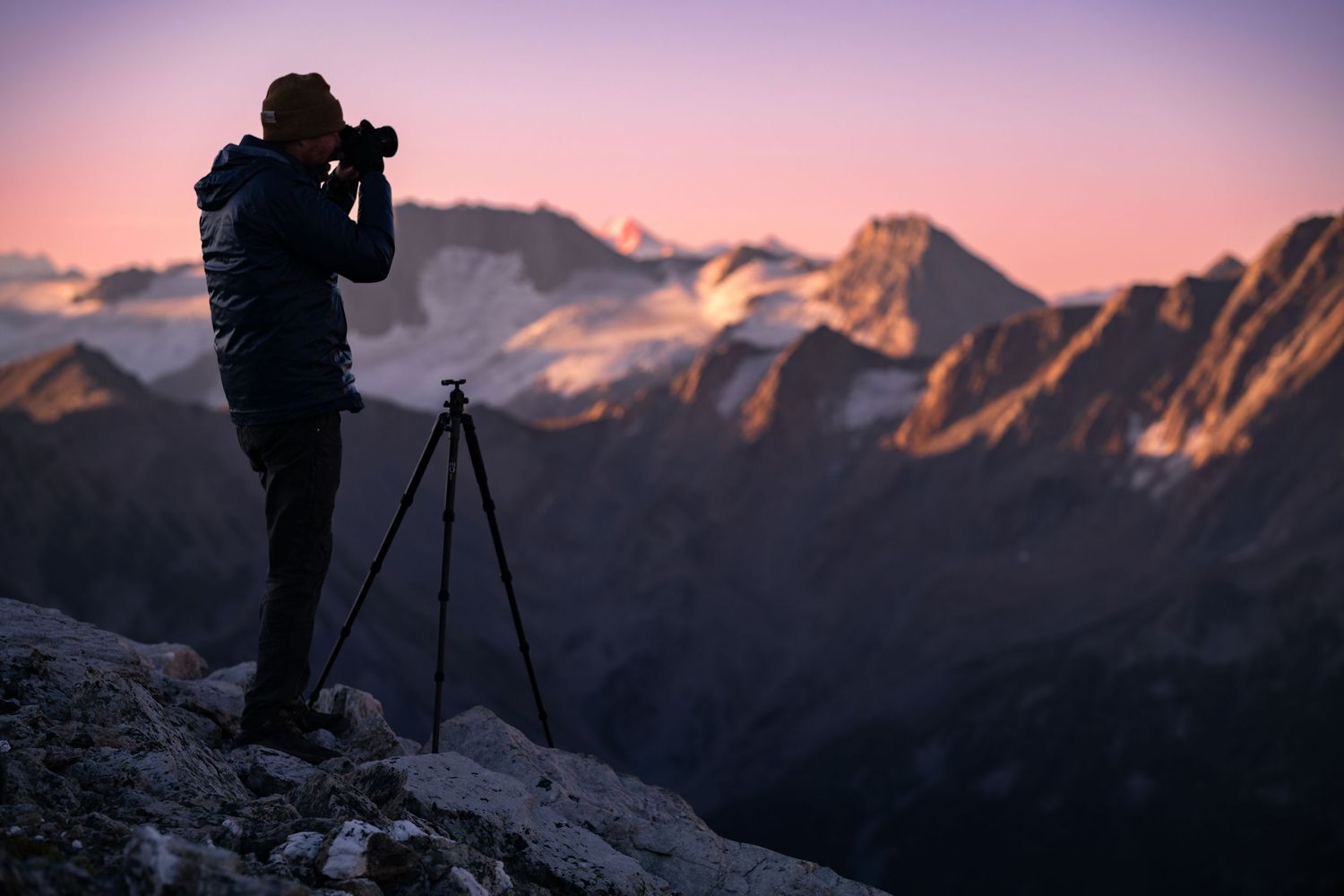 Silhouette of photographer with tripod capturing majestic mountain landscape at twilight with pink and purple sky.