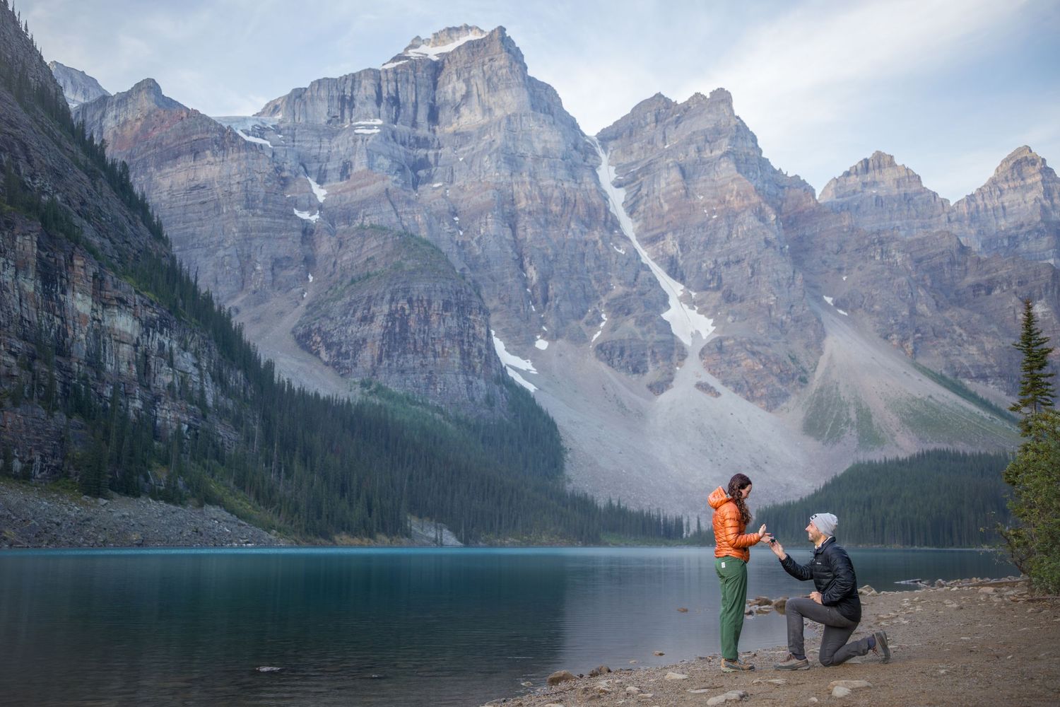 Man proposing to woman by turquoise mountain lake with dramatic rocky peaks in background.