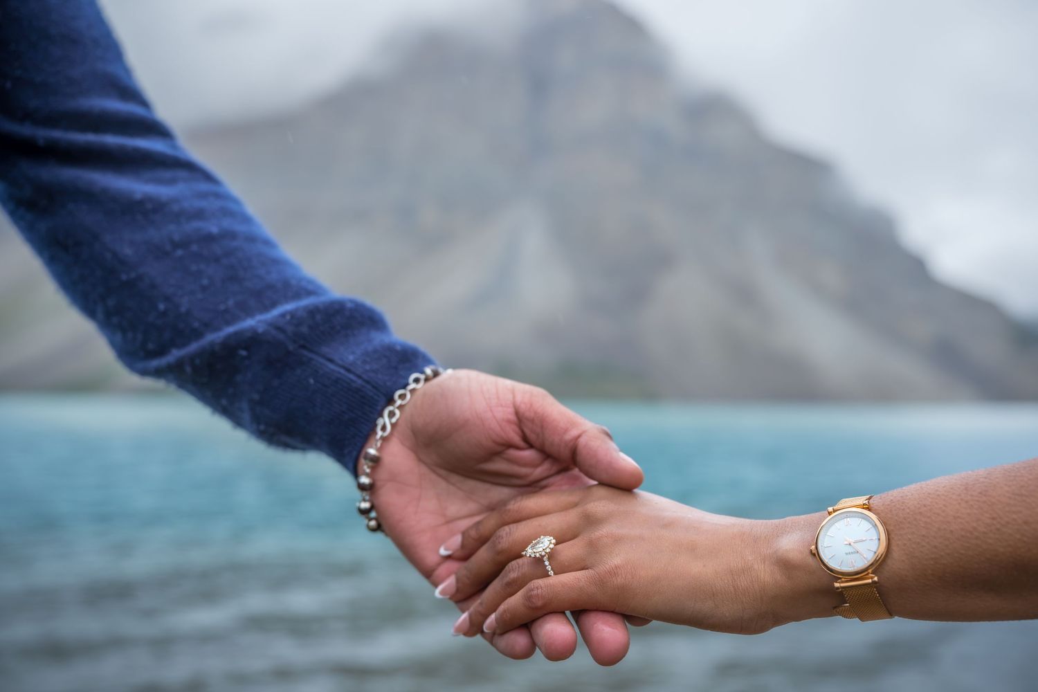 Couple holding hands near a mountain lake, woman wearing a gold watch and engagement ring, man in blue sweater with bracelet.