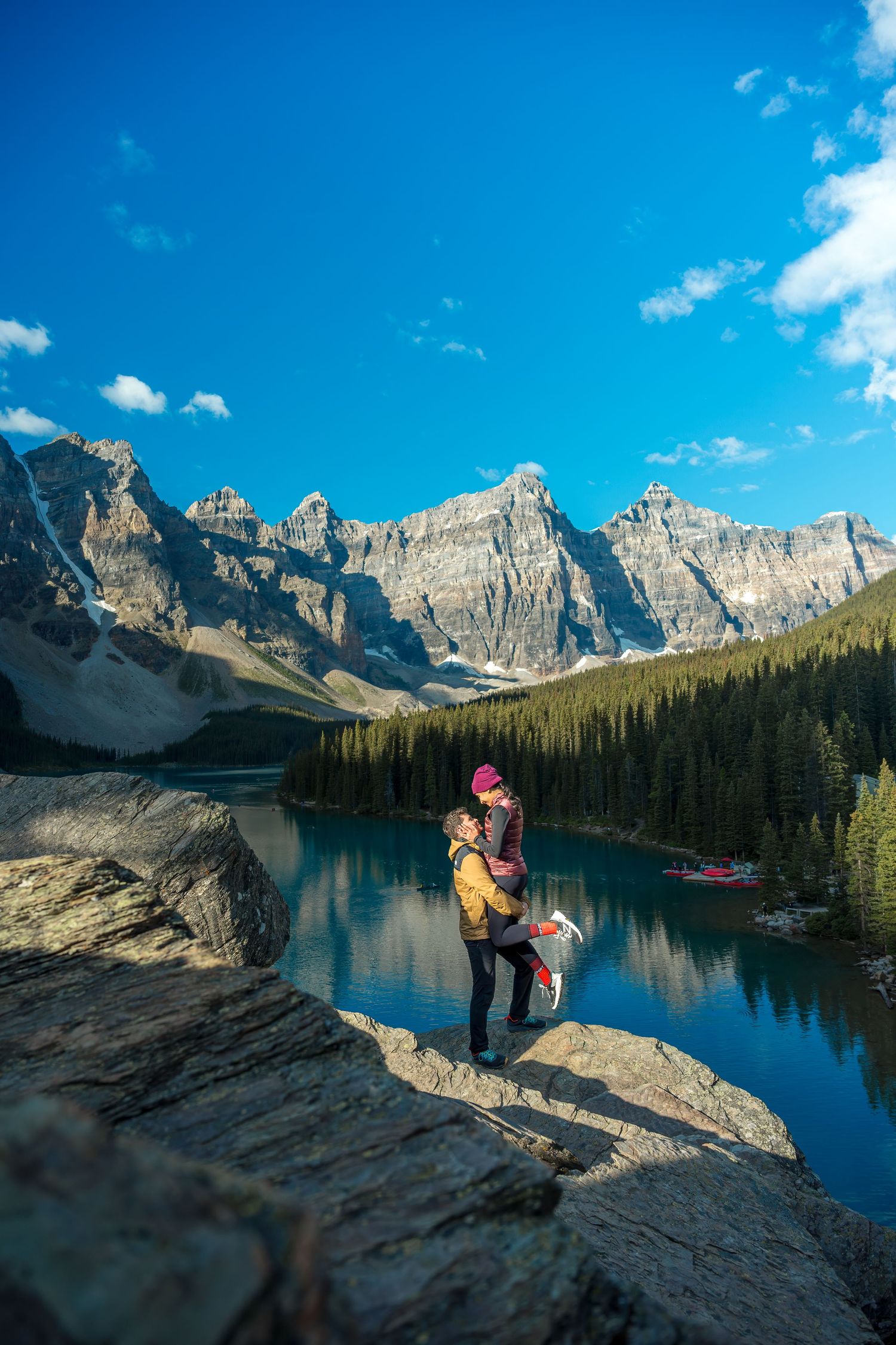 Couple embracing on rocky ledge at Moraine Lake with turquoise water and Canadian Rockies backdrop.